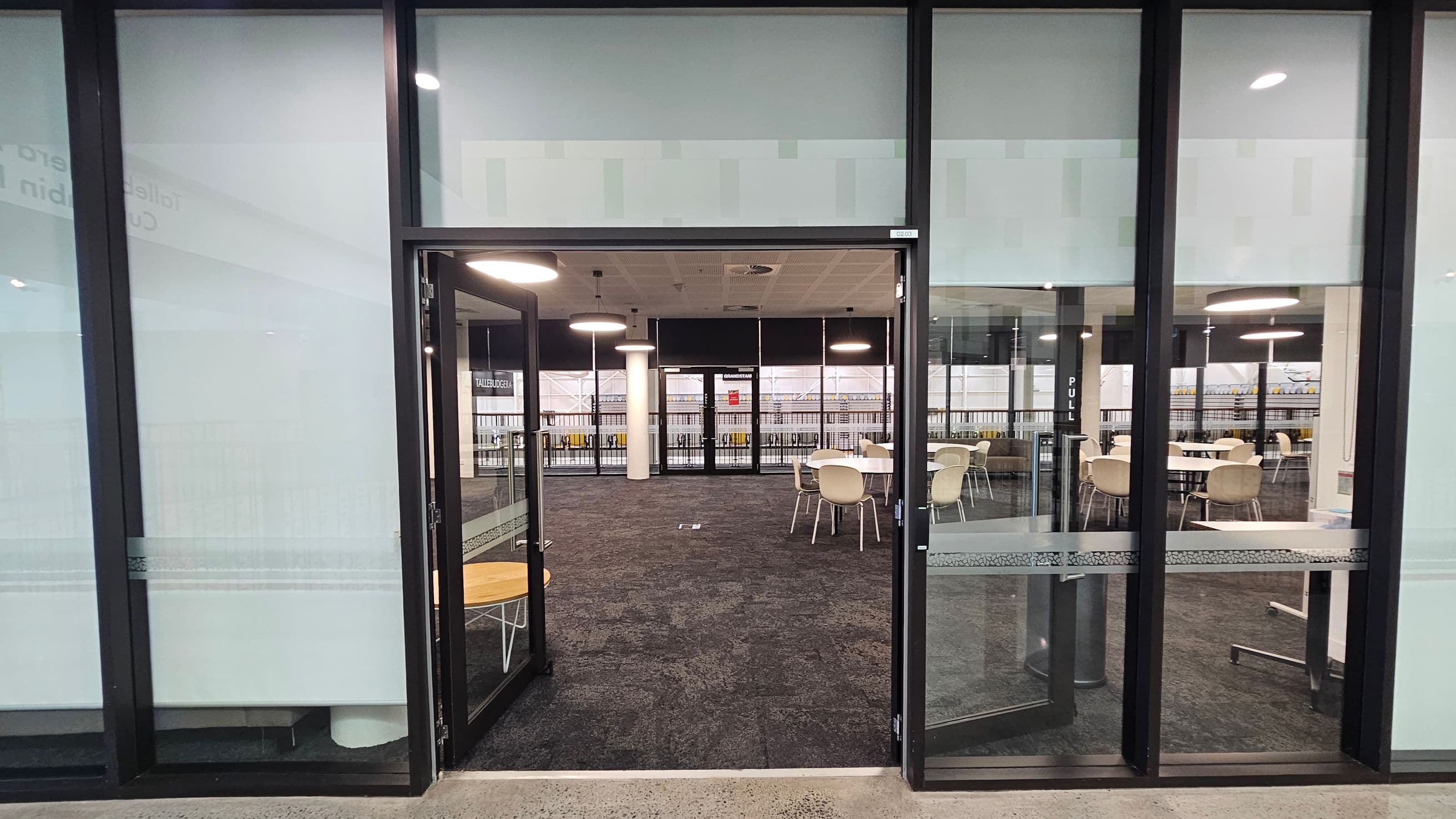 Glass doors open into a room with a dark carpet featuring a subtle pattern. Inside, there are several round tables with white chairs. The ceiling has circular lights, and large windows line the back wall, allowing natural light to enter. The floor outside the room is concrete.