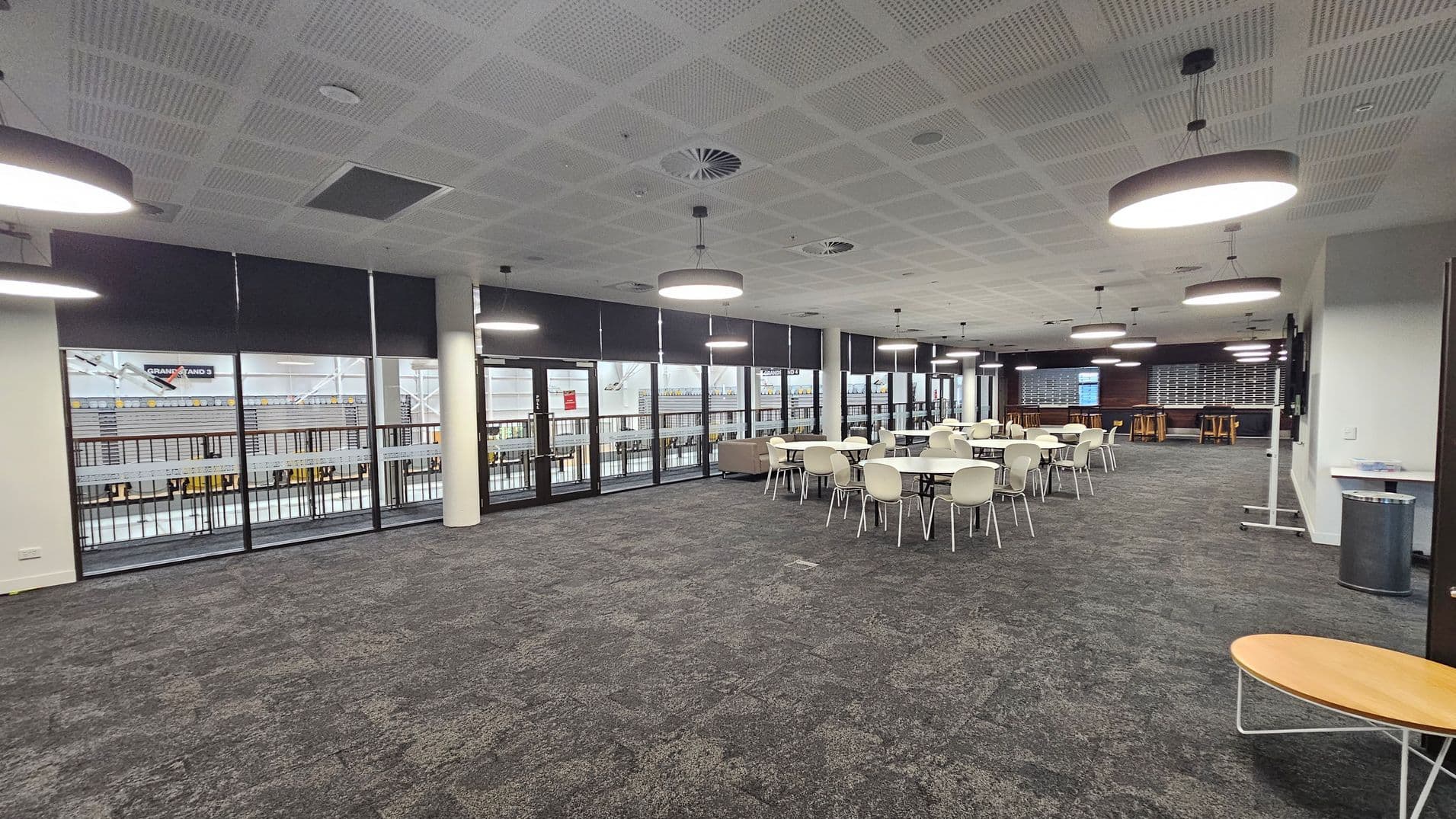 A spacious room with a grey carpeted floor featuring green patches. On the right, there are several round tables with white chairs. In the background, large windows with black blinds overlook an outdoor area. The ceiling is white with circular light fixtures and ventilation panels. On the far right, there's a small round wooden table and a metal bin. The room has a modern, open design with a mix of seating options.