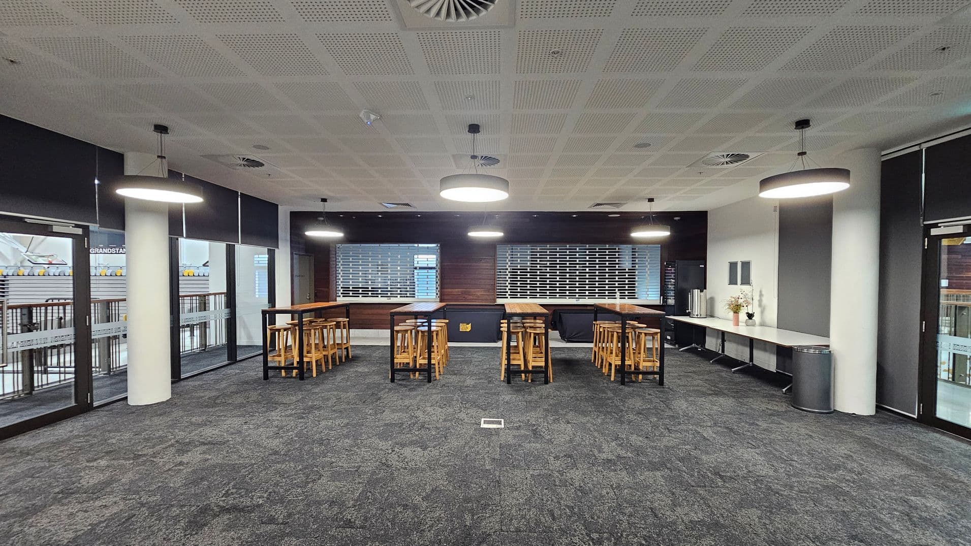 A modern indoor space with a grey carpeted floor and a perforated ceiling with circular lights. In the centre, there are high wooden tables with matching stools. The back wall features two large shuttered windows. On the right, a long counter holds a coffee machine and decorative plants. Large glass doors on the left and right lead to an outdoor area with visible railings.