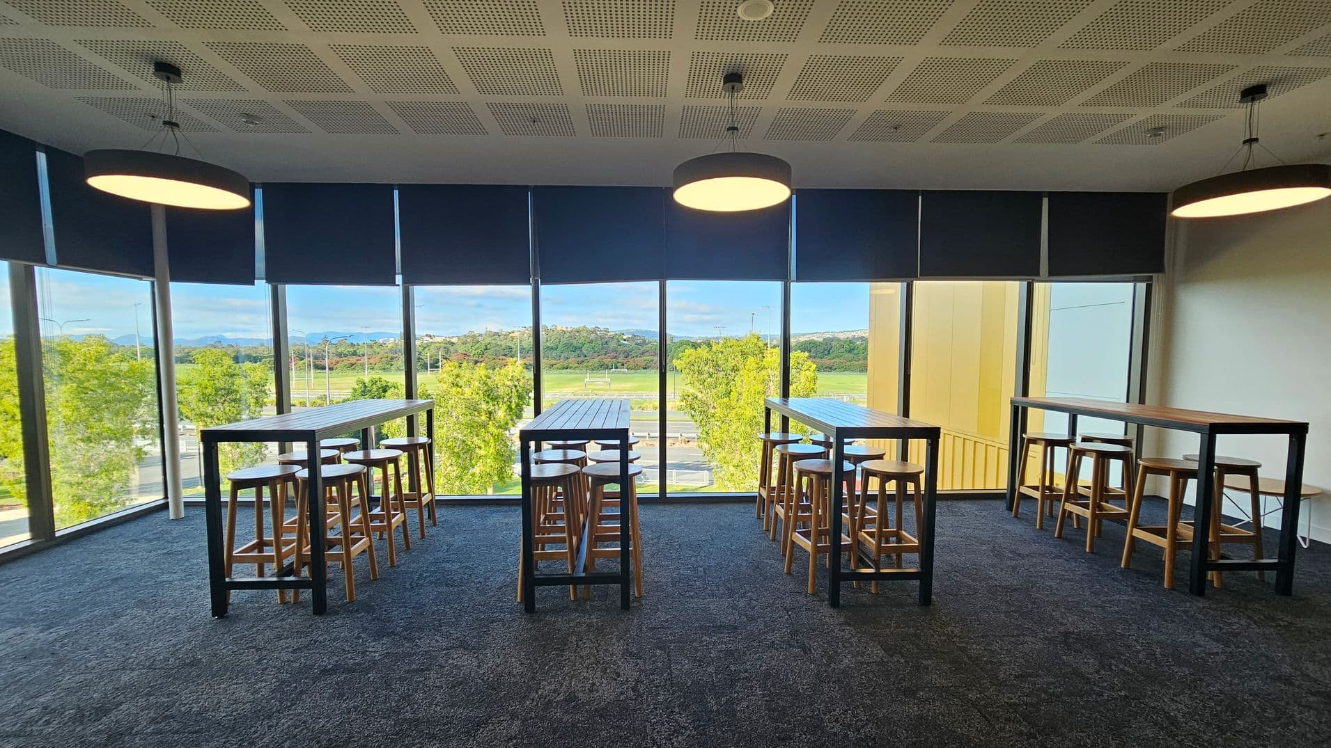 A room with large floor-to-ceiling windows on the left and centre, offering a view of trees and a distant landscape. Three tall tables with wooden stools are aligned in front of the windows. The ceiling is perforated with three round pendant lights hanging above. The floor is carpeted in a dark colour. The right side of the room has a yellow wall panel.