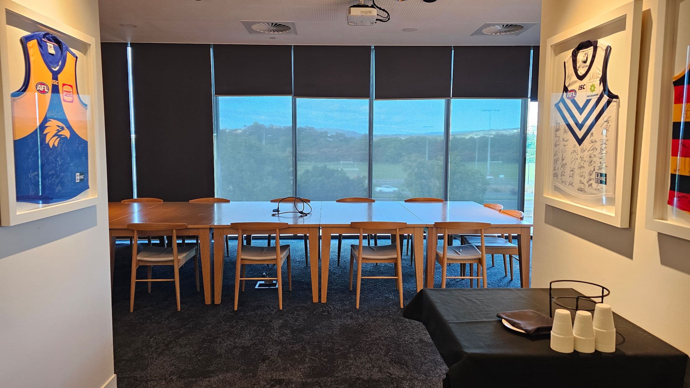 A meeting room with a long wooden table and several chairs in the centre. The back wall features large windows with blinds partially drawn, revealing a view of greenery outside. On the left and right walls, framed sports jerseys are displayed. In the foreground, a small table is covered with a black cloth, holding a stack of white cups and a metal holder. The floor is carpeted, and the ceiling has recessed lighting and a projector.