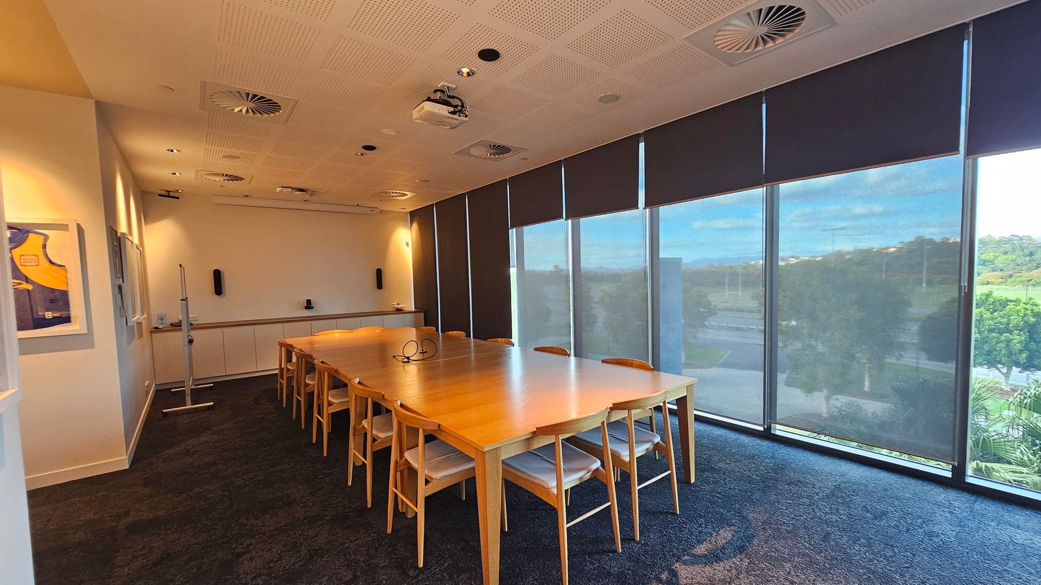 A conference room with a large wooden table surrounded by wooden chairs with white cushions. On the left wall, there are framed pictures, including a sports jersey. The right side features floor-to-ceiling windows with dark blinds partially drawn, offering a view of greenery outside. The ceiling has recessed lighting and air vents, and a projector is mounted above the table. The floor is carpeted in a dark colour.