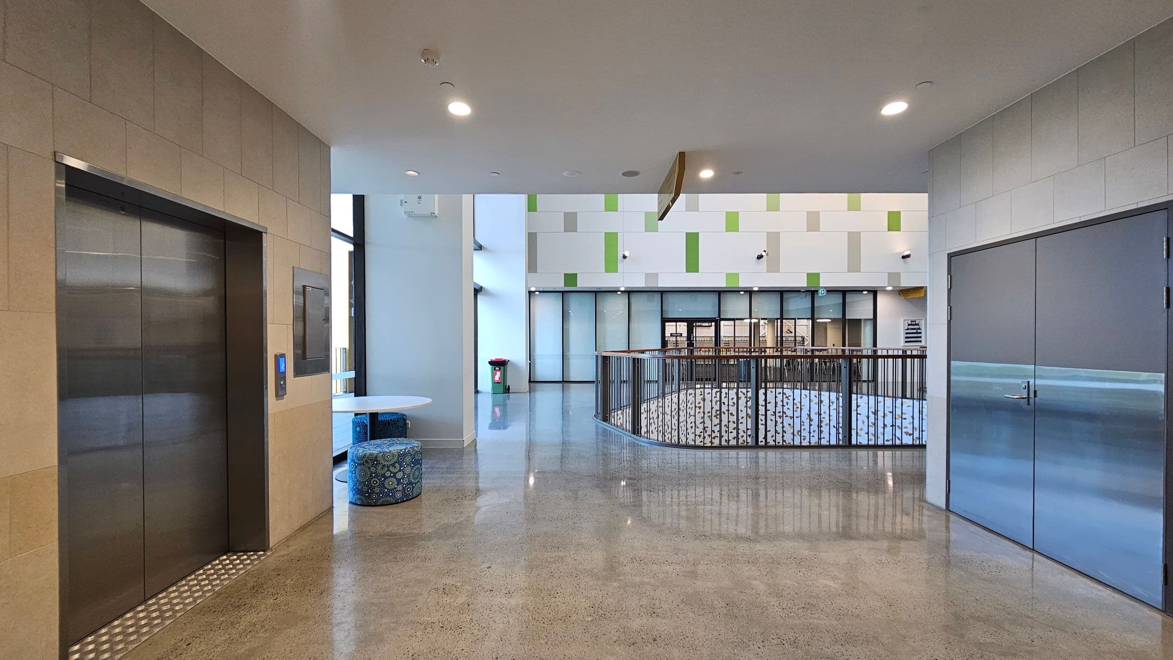 A modern interior space with a polished concrete floor. On the left, there is a stainless steel lift with beige tiled walls. Near the lift, a small round table with patterned blue stools is visible. In the centre, a railing surrounds an open area below. The back wall features a geometric pattern in white and green, with large glass doors. On the right, there are double doors with a metallic finish. The ceiling is white with recessed lighting.