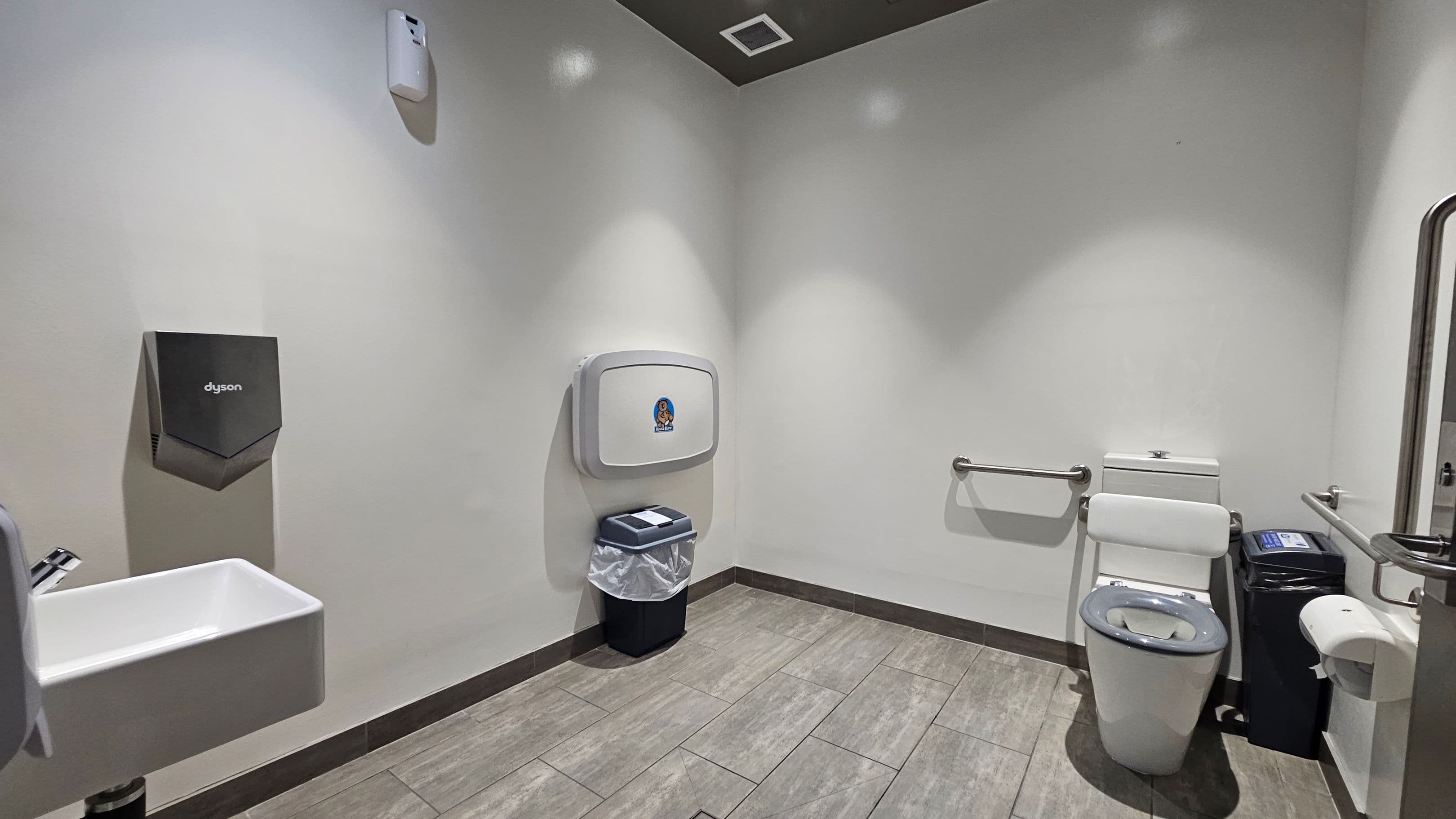 A public restroom with grey tiled flooring and white walls. On the left, a Dyson hand dryer is mounted above a small white sink. In the centre, a baby changing station is above a bin. On the right, a toilet with a safety rail is next to a small bin and a toilet paper dispenser. The ceiling is dark with recessed lighting and a vent.