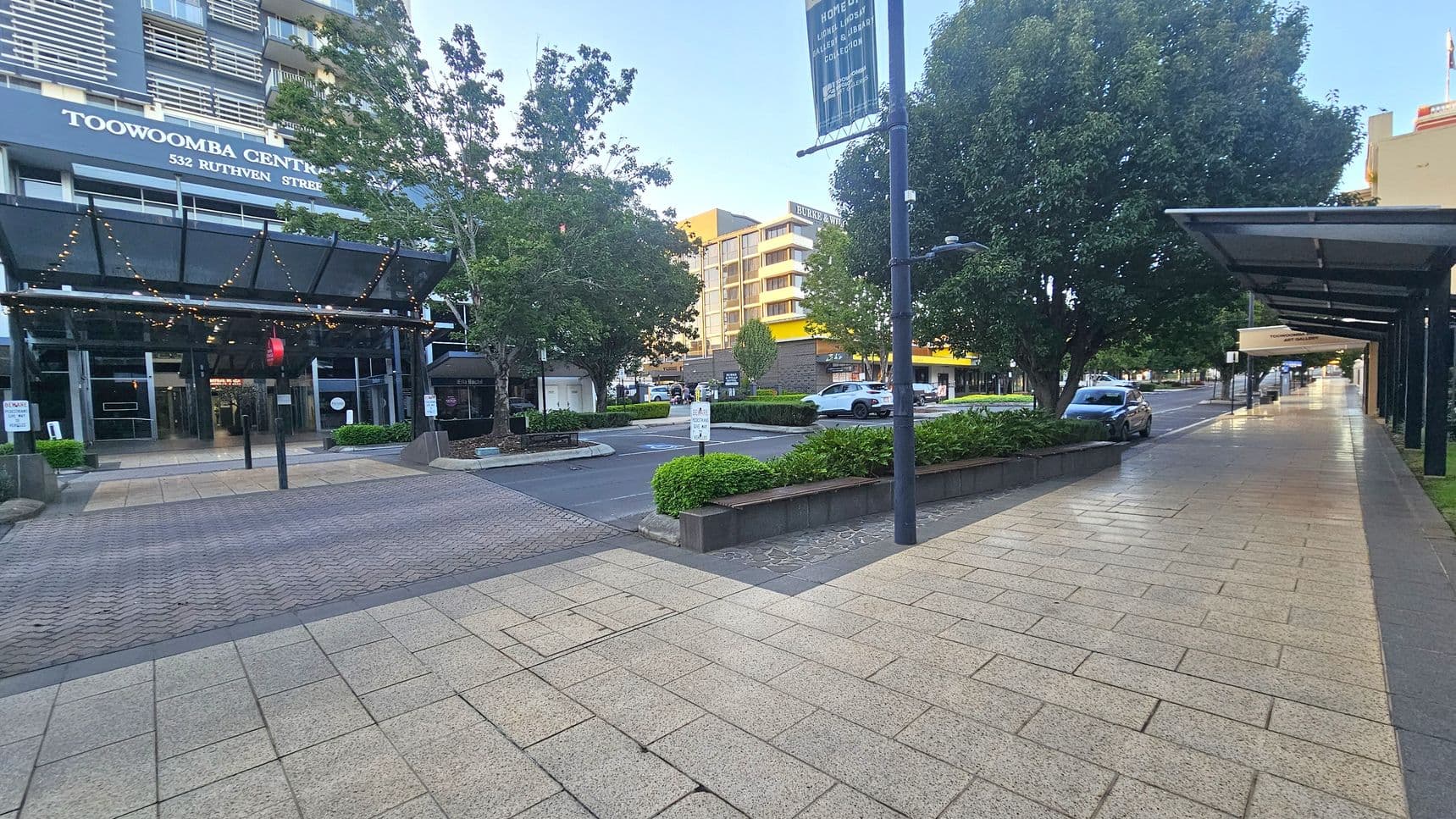 A city street scene with a building on the left labelled "Toowoomba Centre, 532 Ruthven Street". The entrance is covered by a glass canopy adorned with string lights. Trees and planters line the street, with a few parked cars visible. On the right, a long pavement stretches under a series of covered walkways. The ground is paved with light-coloured tiles, and the area appears clean and well-maintained.