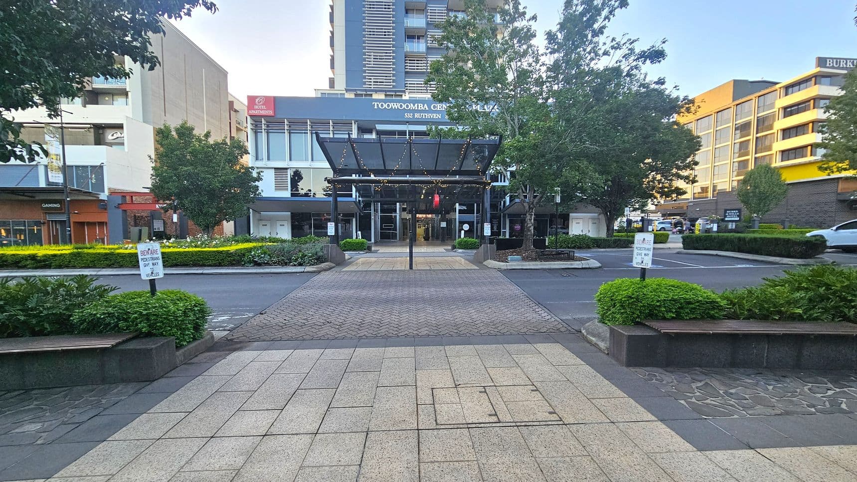 A paved walkway leads to a building with a glass canopy adorned with lights. The building has signage for "Toowoomba Central Plaza" and "Hotel Apartments." Trees and shrubs line the path, with a "Beware Pedestrians Give Way to Vehicles" sign visible. On the left, there's a gaming establishment, while the right features a multi-storey building with yellow accents. The foreground has a mix of stone and tile paving.