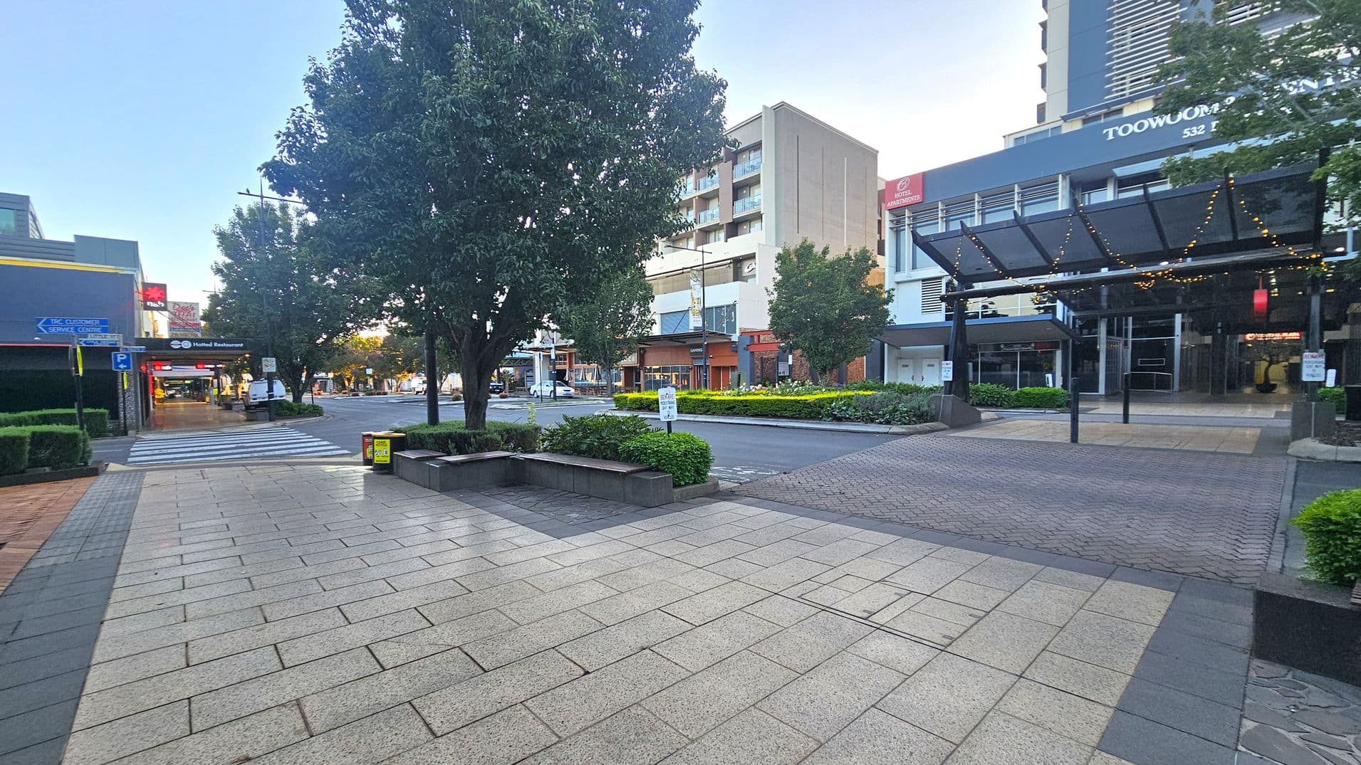 A city street scene with a paved pedestrian area in the foreground. On the left, there are shops with signs and a zebra crossing leading to a tree-lined street. In the centre, a large tree and some greenery are visible. On the right, a modern building with a glass canopy and string lights is present. The background features more buildings, including a tall structure with balconies. The area appears clean and well-maintained.