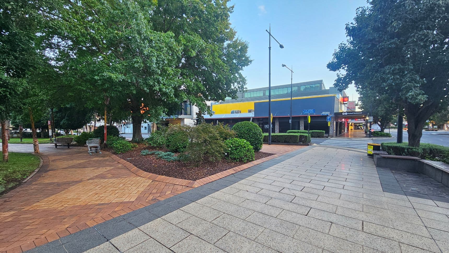 A paved walkway with light grey tiles on the right and reddish-brown bricks on the left. On the left, there are large trees and a bench with a shopping trolley nearby. In the centre, there are neatly trimmed bushes and a small garden area. On the right, a modern building with a yellow and blue facade is visible, along with a zebra crossing and street lamps. The area is surrounded by more trees and urban structures.
