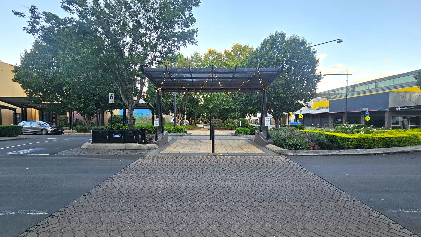 A paved walkway leads to a pergola adorned with string lights in the centre. Behind it, a park area with trees and shrubs is visible. On the left, a parked car is near a building with a mural. On the right, a building with a yellow and grey facade is adjacent to a garden with yellow and green plants. The foreground features a patterned brick pavement, and the background is filled with trees.