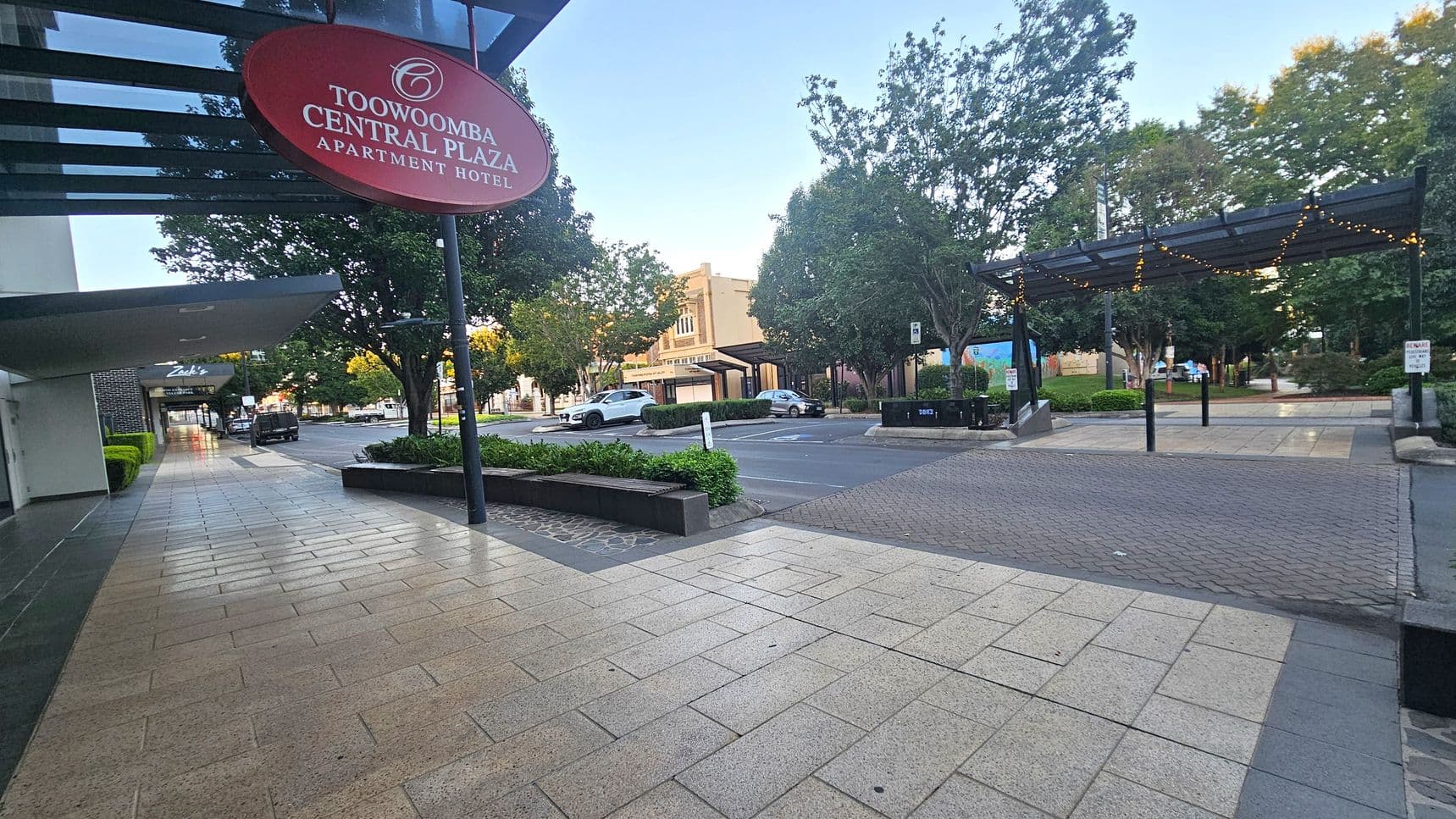 A street scene with a wide pavement on the left, featuring a red sign for "Toowoomba Central Plaza Apartment Hotel." The pavement is tiled in light and dark grey. Across the street, there are trees and a small park area with a covered walkway adorned with string lights. A few cars are parked along the street, and a building with a beige facade is visible in the background. The area is lined with greenery and benches.