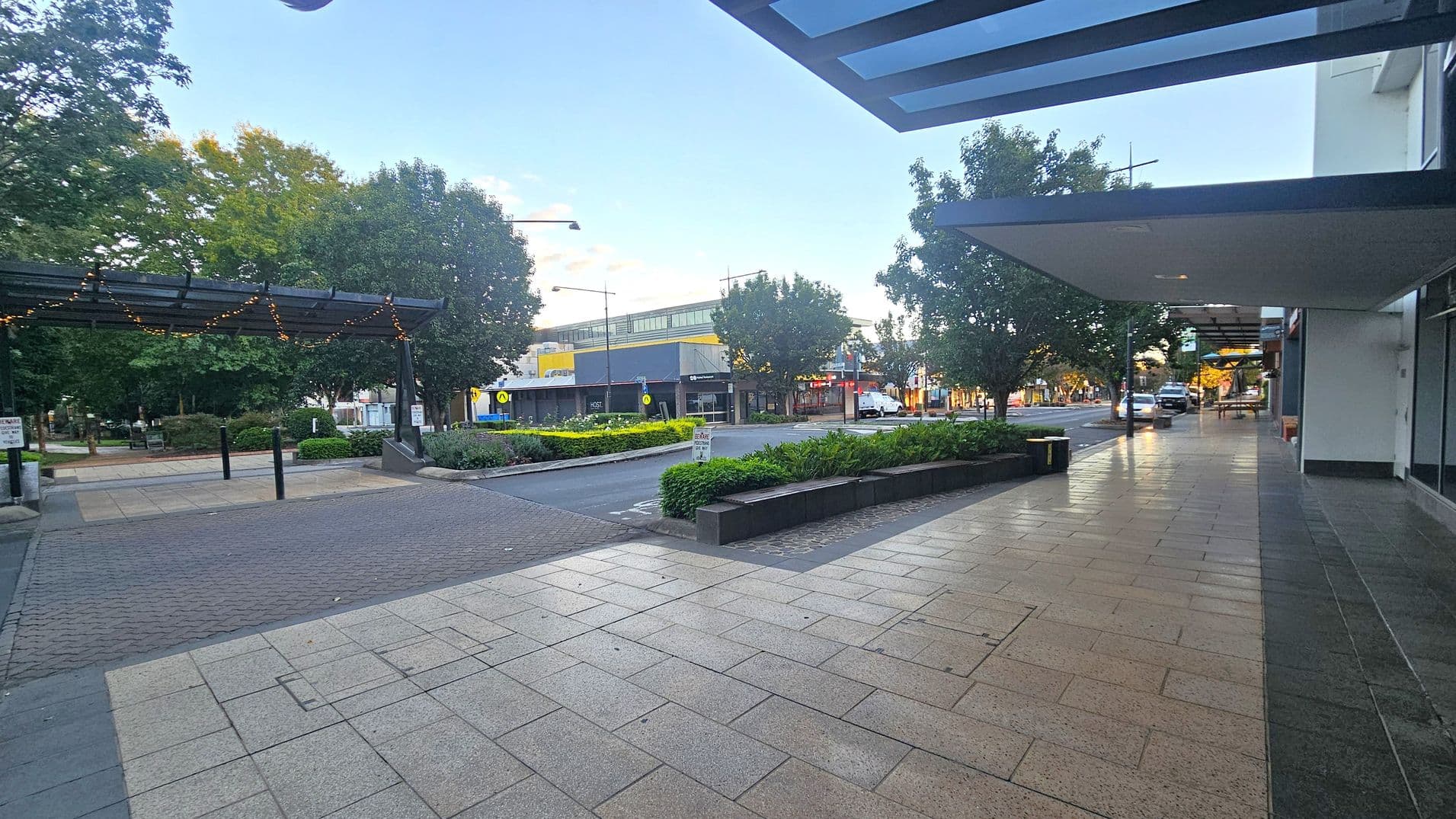 A street scene with a paved walkway on the right, bordered by a building with a glass awning. Trees line the street, providing shade. On the left, a small park area with a pergola adorned with string lights. In the background, a road with a few vehicles and a building with a yellow and grey facade. The pavement is a mix of light and dark tiles, and there are planters with greenery along the walkway.