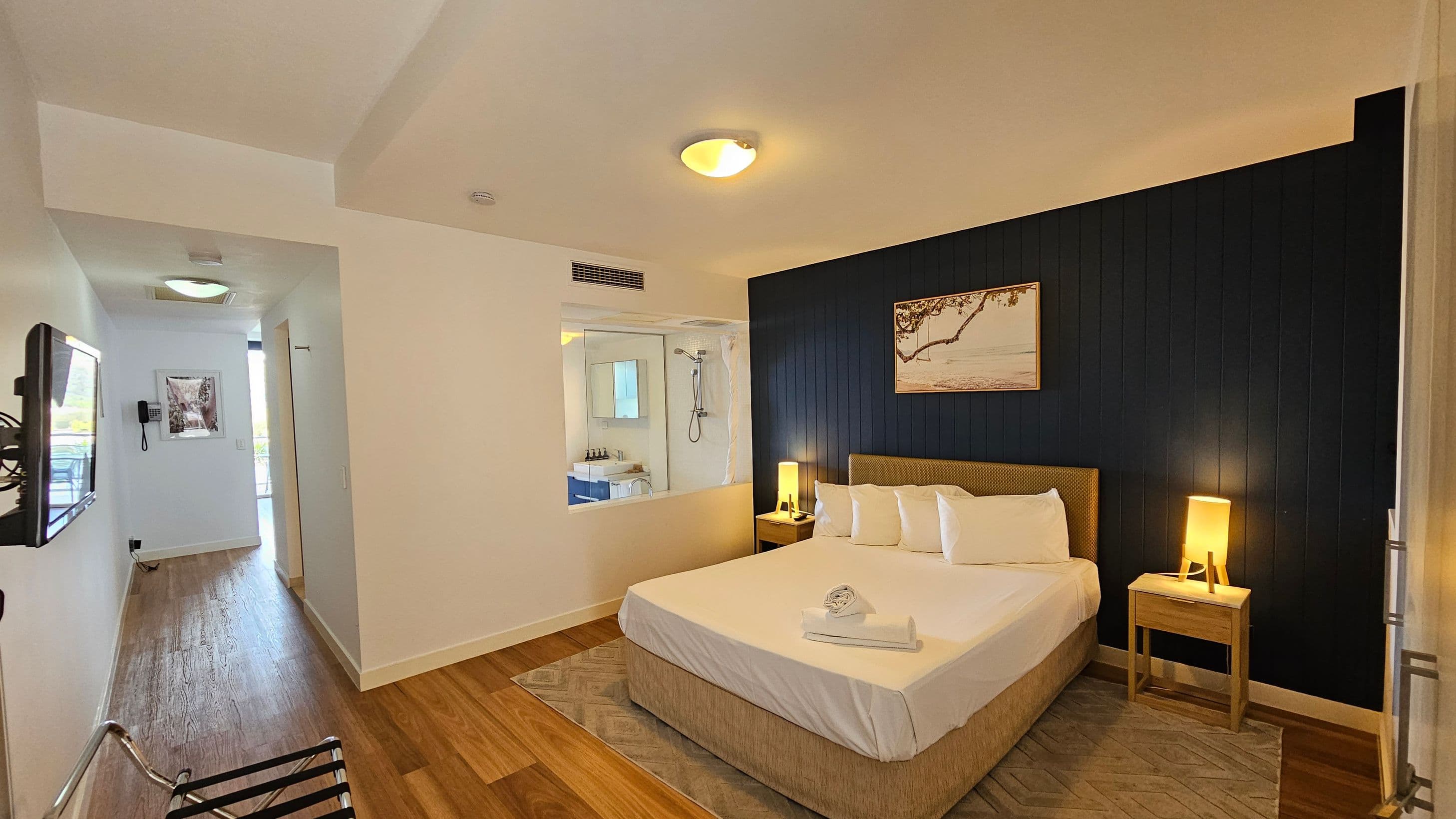 A hotel room with a large bed in the centre, topped with white linens and towels. On either side of the bed are wooden bedside tables with lamps. The wall behind the bed is dark with a framed picture. To the left, a hallway with wooden flooring leads to a bathroom visible through a glass partition. A wall-mounted TV and a telephone are on the left wall. The ceiling is light-coloured with recessed lighting.