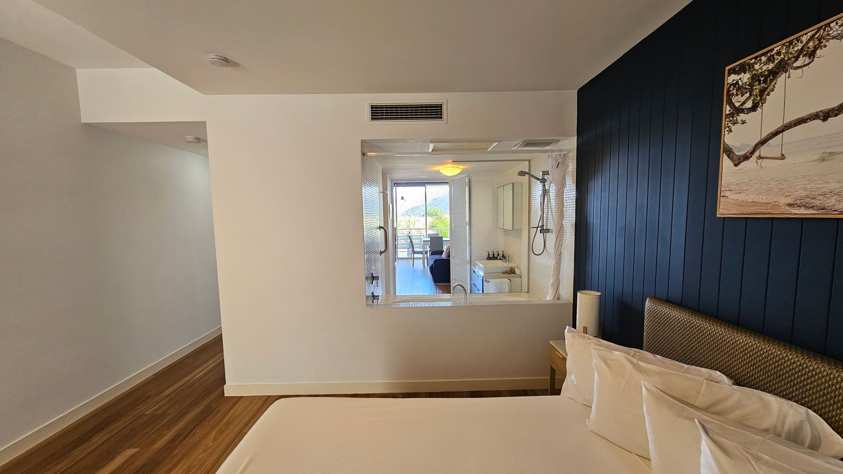 A modern bedroom with a wooden floor and a white ceiling. On the right, there's a bed with white pillows and a woven headboard against a dark blue panelled wall. A framed picture of a tree with a swing hangs above. In the centre, a large window reveals a bathroom with a shower and sink, leading to a balcony with a view of greenery and distant hills. On the left, a narrow hallway is visible.