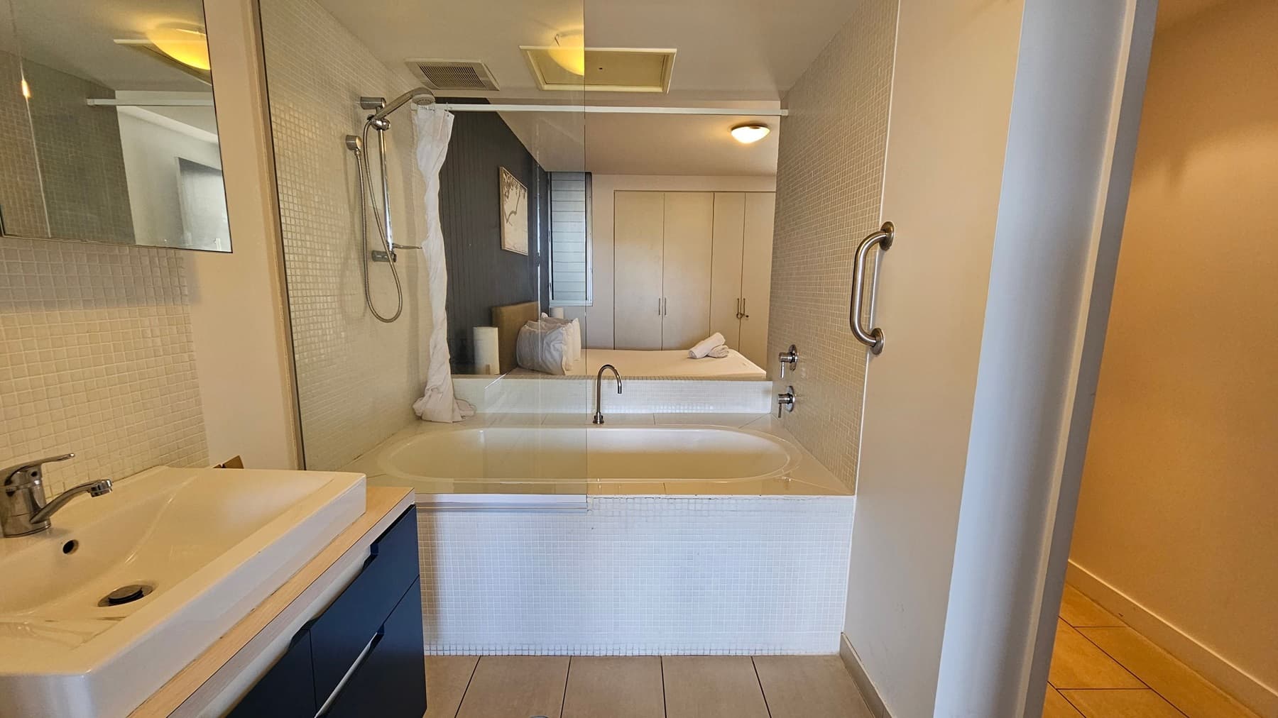 A bathroom with a white sink and chrome tap on the left, set on a light-coloured countertop with dark cabinets below. The wall is covered in small white tiles. In the centre, there's a bathtub with a showerhead above and a white curtain partially drawn. A large mirror reflects a bedroom with a bed, pillows, and a wardrobe. The floor is tiled in a light beige colour, and a metal grab bar is mounted on the right wall.