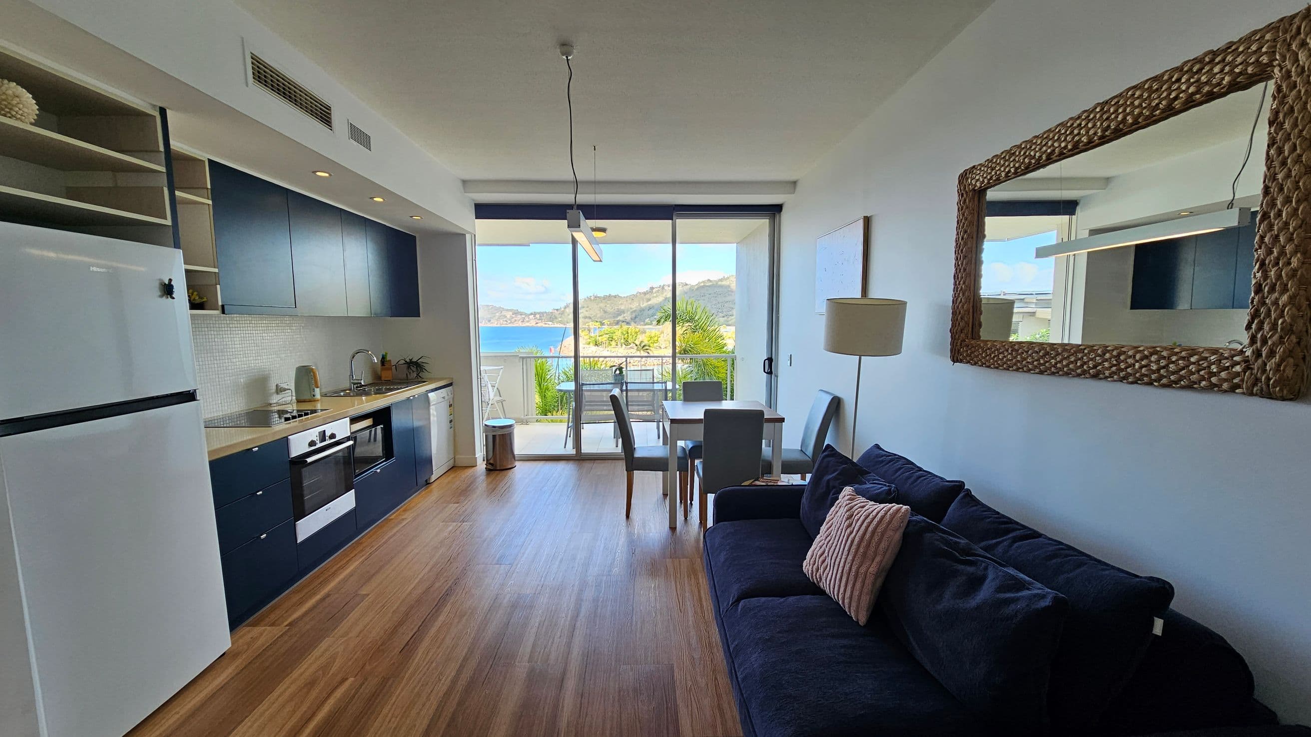 A modern living space with a kitchen on the left featuring blue cabinets, a white fridge, and a built-in oven. The floor is wooden. In the centre, a dining table with chairs is positioned near large glass doors leading to a balcony with a view of the sea and hills. On the right, a dark sofa with a cushion is placed under a large woven-framed mirror. A floor lamp stands nearby. The ceiling has recessed lighting and a hanging light fixture.