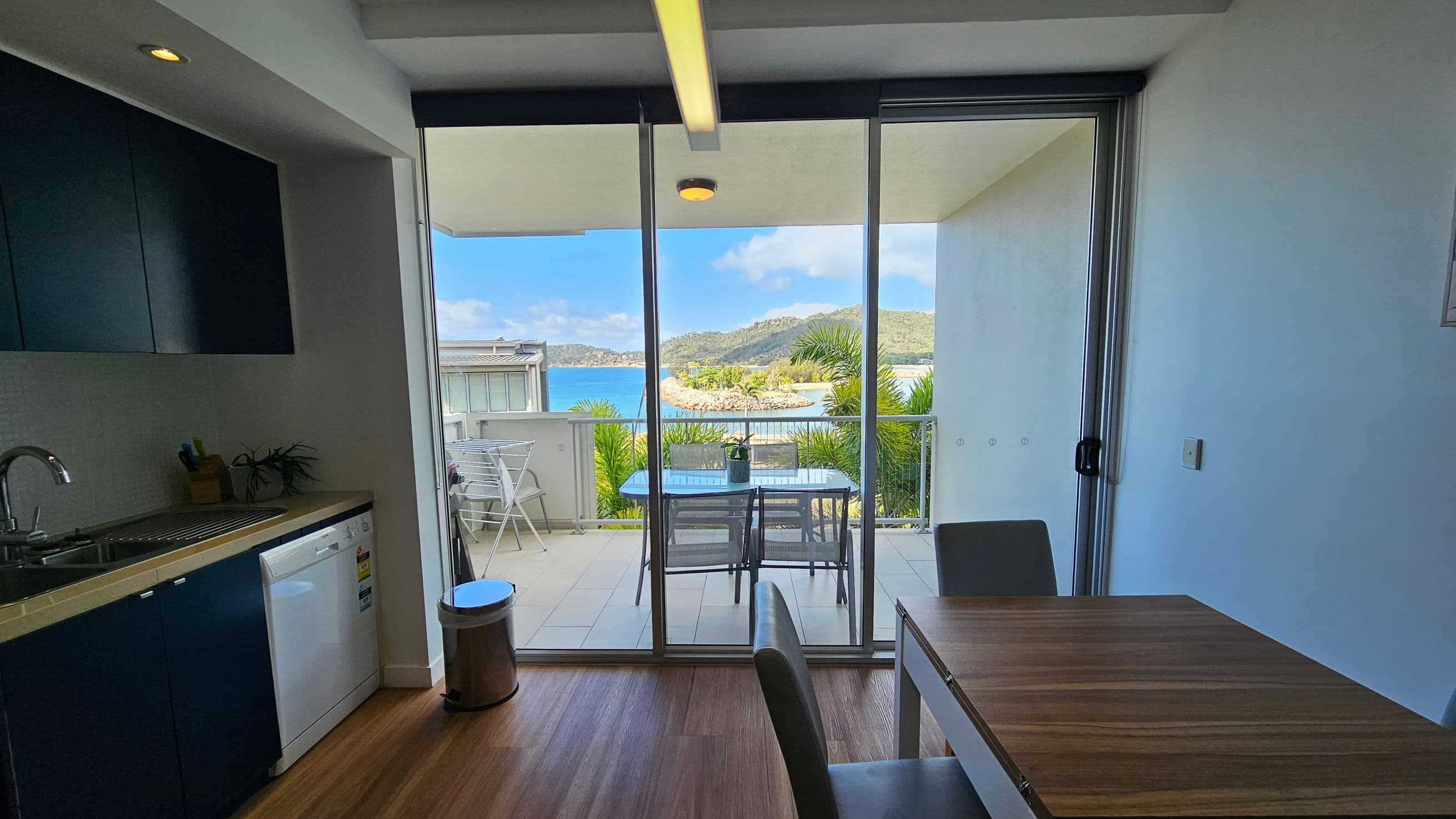 A modern kitchen and dining area with wooden flooring. On the left, there is a kitchen counter with a sink, a dishwasher, and a small plant. A metal bin is nearby. In the centre, large glass doors open to a balcony with a table and chairs, overlooking a scenic view of the sea and distant hills. On the right, a wooden dining table with chairs is visible. The ceiling has recessed lighting.