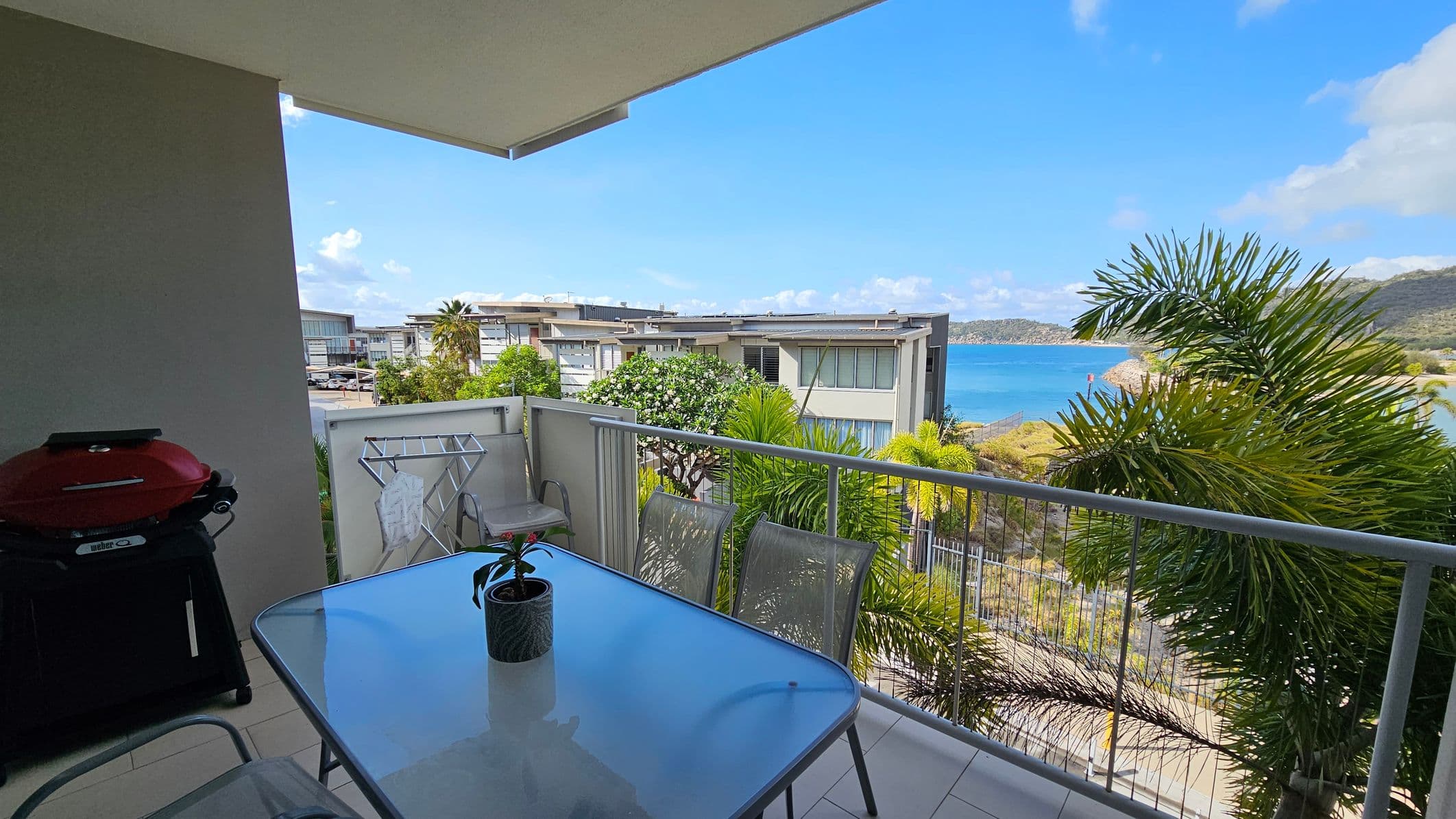 A balcony with a glass table and four chairs, one of which has a small potted plant. On the left, there's a red barbecue grill. A clothes drying rack is next to the railing. In the background, modern buildings are surrounded by lush greenery, and beyond them, a view of the sea and distant hills. The floor is tiled, and the ceiling is a simple overhang.