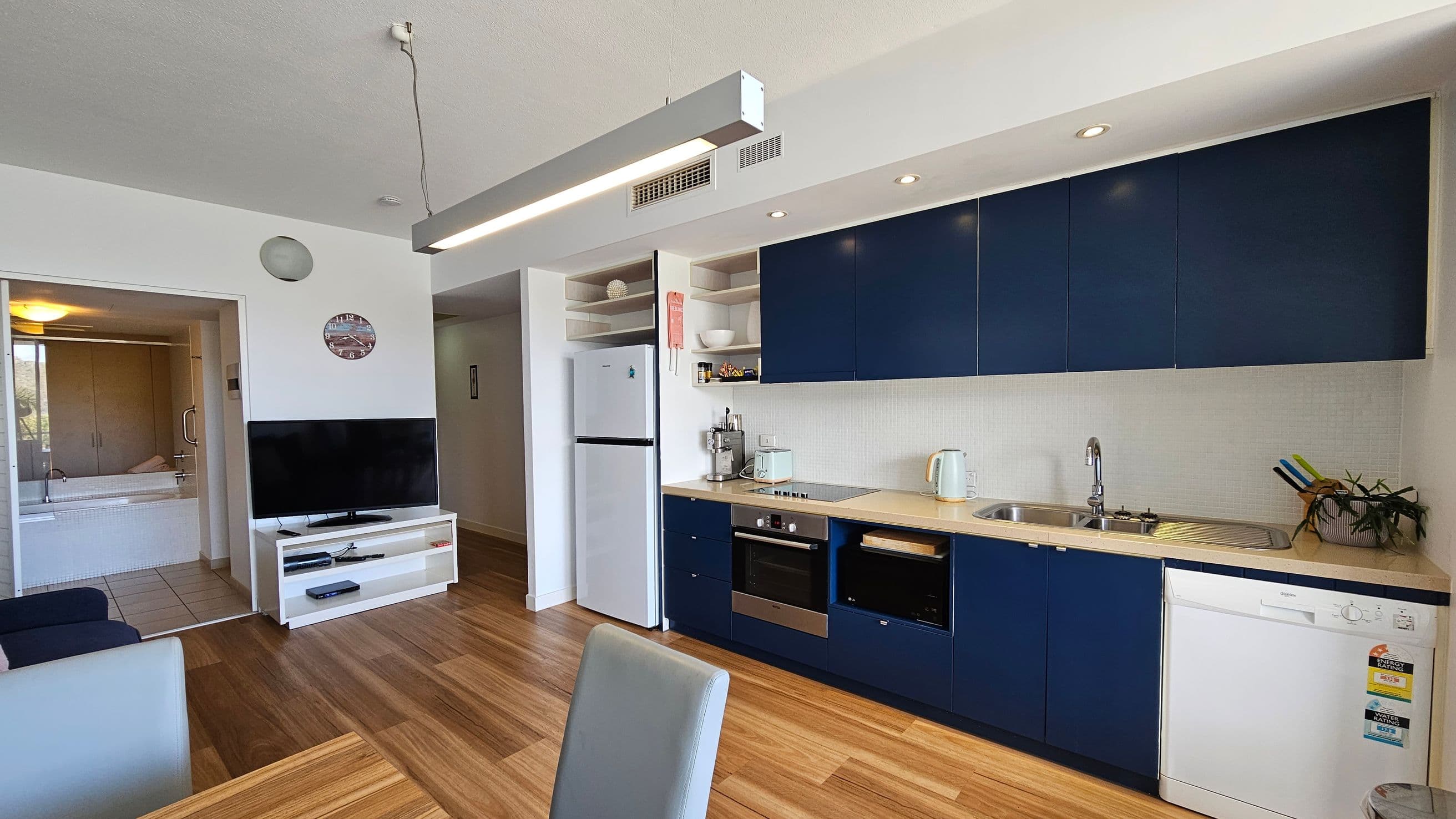 A modern kitchen and living area with wooden flooring. On the right, there are dark blue cabinets, a sink, a kettle, and a dishwasher. A white fridge is next to open shelves. In the centre, a dining chair is visible. On the left, a TV sits on a white stand, and a glimpse of a bathroom with a bathtub is seen through an open door. The ceiling has recessed lighting and a hanging light fixture.