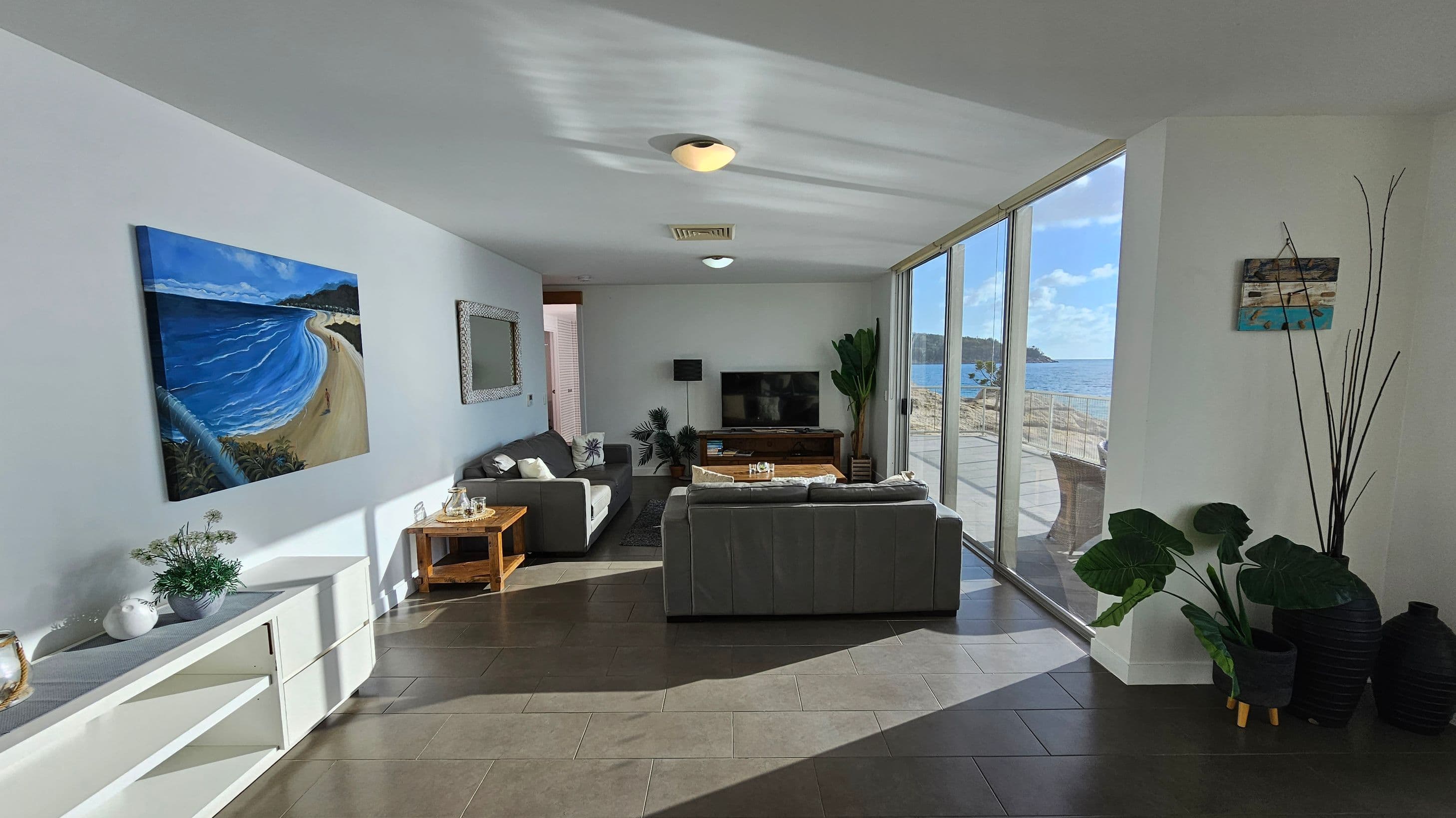 A modern living and dining area with large floor-to-ceiling windows on the right, offering a view of the ocean. The dining area features a wooden table with white chairs. The living area on the left has grey sofas, a TV, and decorative plants. Artwork hangs on the walls, and a lamp sits on a side table. The floor is tiled, and the ceiling has recessed lighting.