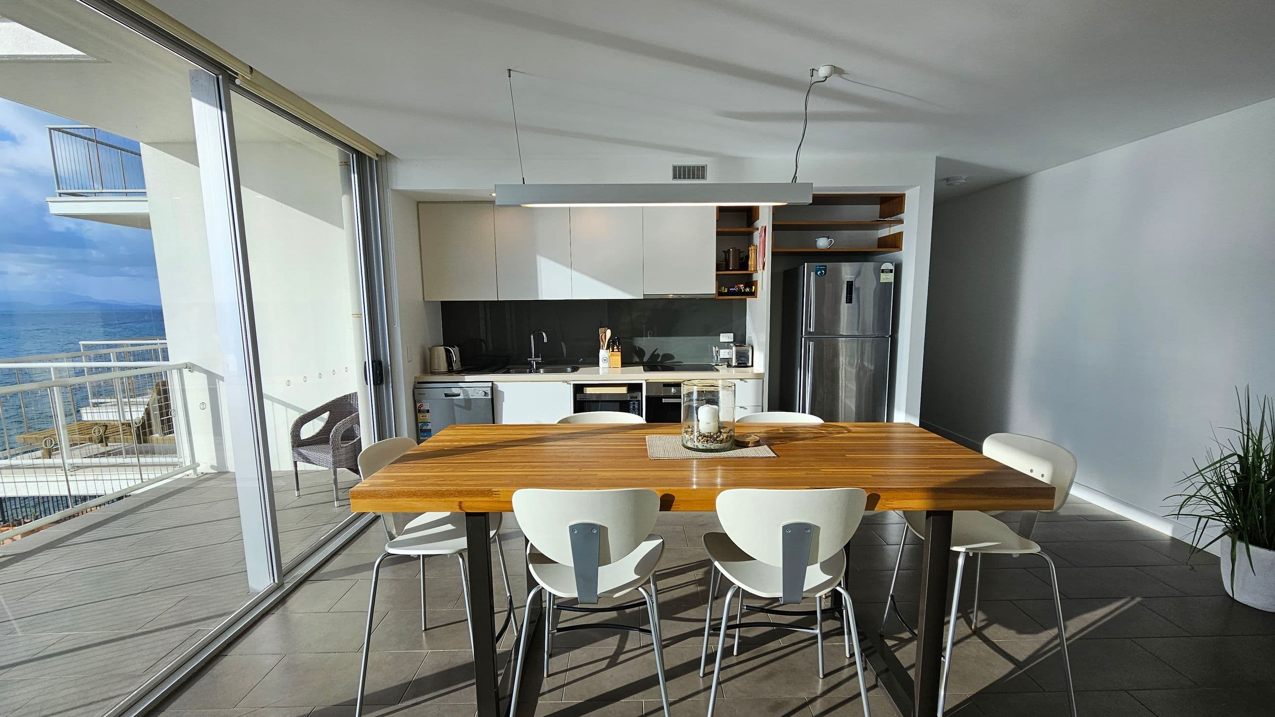 A modern kitchen with a wooden dining table in the centre, surrounded by six white chairs. The background features a sleek kitchen area with white cabinets, a black backsplash, and stainless steel appliances, including a fridge. To the left, large glass sliding doors open to a balcony with a view of the sea. The floor is tiled in a dark grey, and a potted plant is in the right corner. The ceiling is white with a minimalist light fixture above the table.