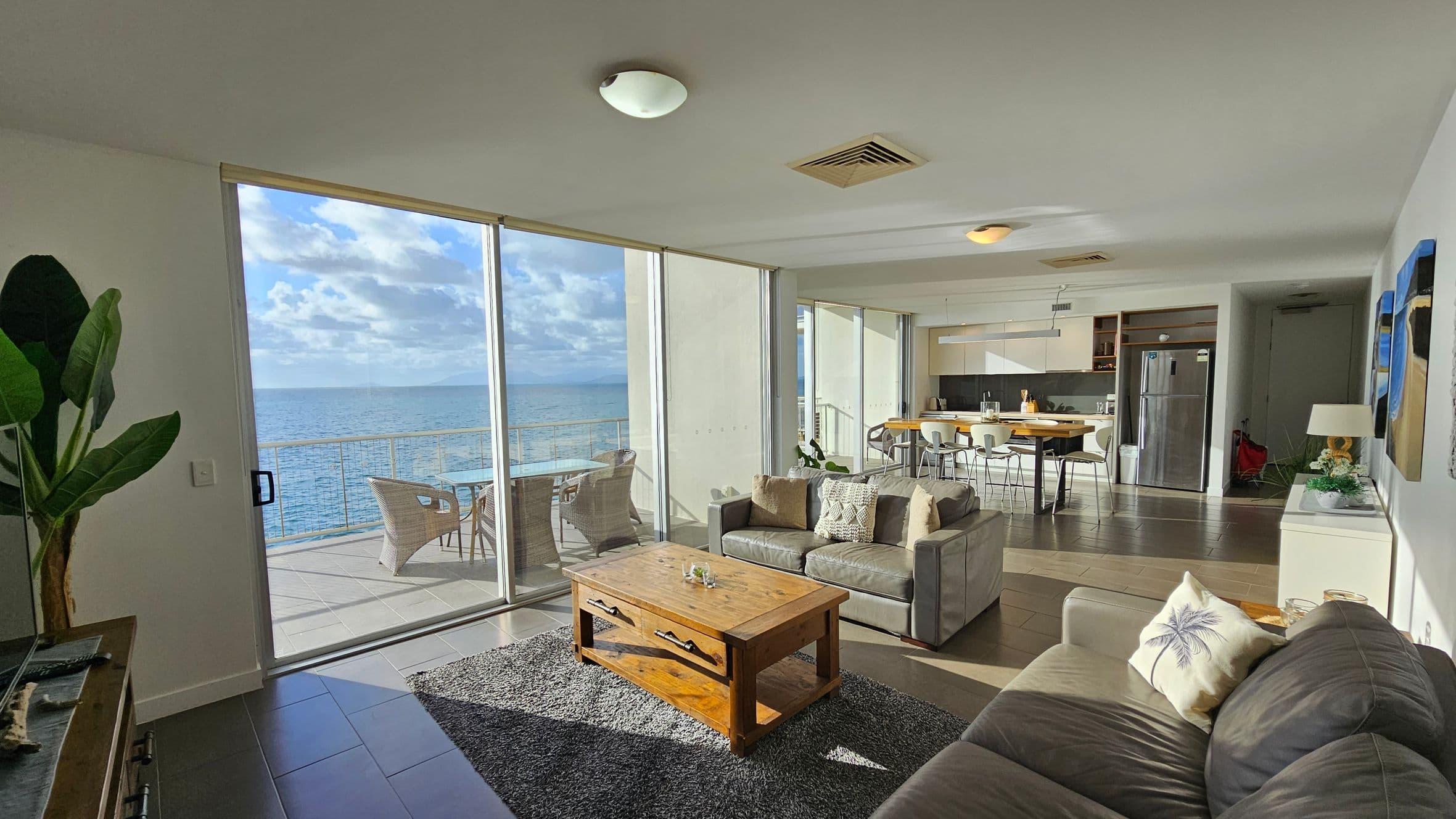 A modern living room with large glass sliding doors on the left, offering a view of the ocean and a balcony with wicker chairs. In the centre, a grey sofa with cushions faces a wooden coffee table on a grey rug. The right side features a dining area with a table and chairs, and a kitchen with a fridge and cabinets. The ceiling has recessed lighting, and a plant is visible in the foreground. The floor is tiled, and there are decorative items on a side table.