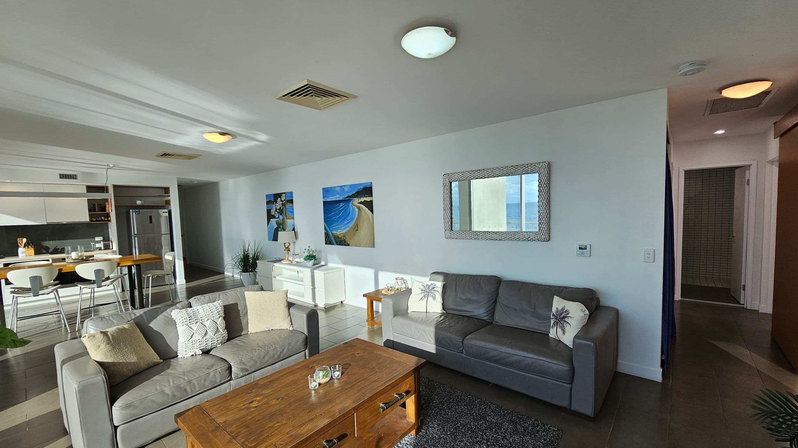 A modern living room with two grey sofas, each adorned with decorative cushions, facing a wooden coffee table. On the wall behind the sofas, there's a large mirror and two beach-themed paintings. To the left, a dining area with a wooden table and white chairs is visible, along with a kitchen featuring a stainless steel fridge. The floor is tiled, and the ceiling has recessed lighting and air vents. A hallway leads to another room on the right.