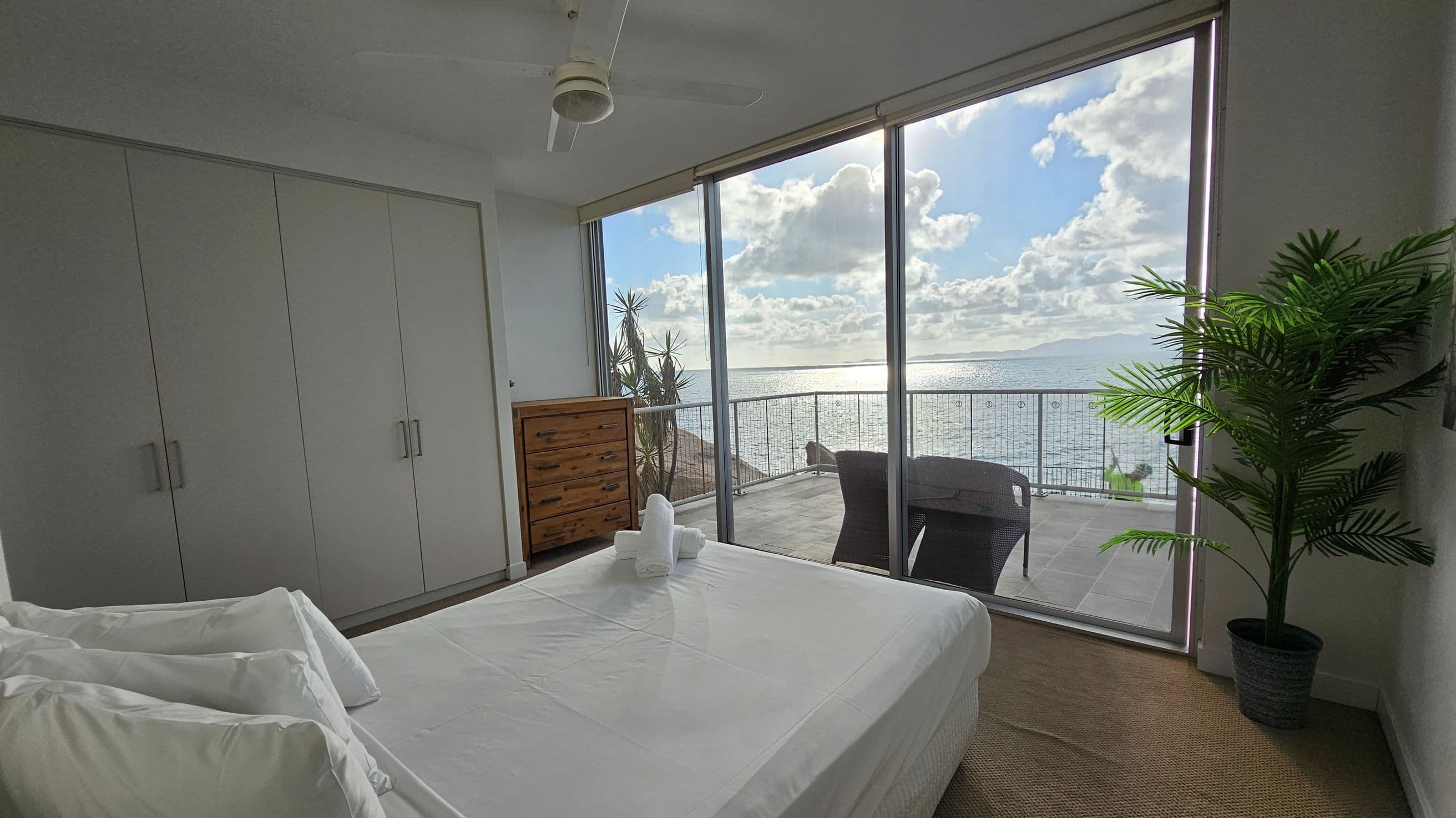 A bedroom with a large window overlooking the sea. On the left, there are white wardrobes and a wooden chest of drawers. In the centre, a neatly made bed with white linens and towels is positioned. To the right, a potted plant adds greenery. Outside, a balcony with a chair offers a view of the ocean. The floor is covered with a woven rug, and a ceiling fan is visible above.
