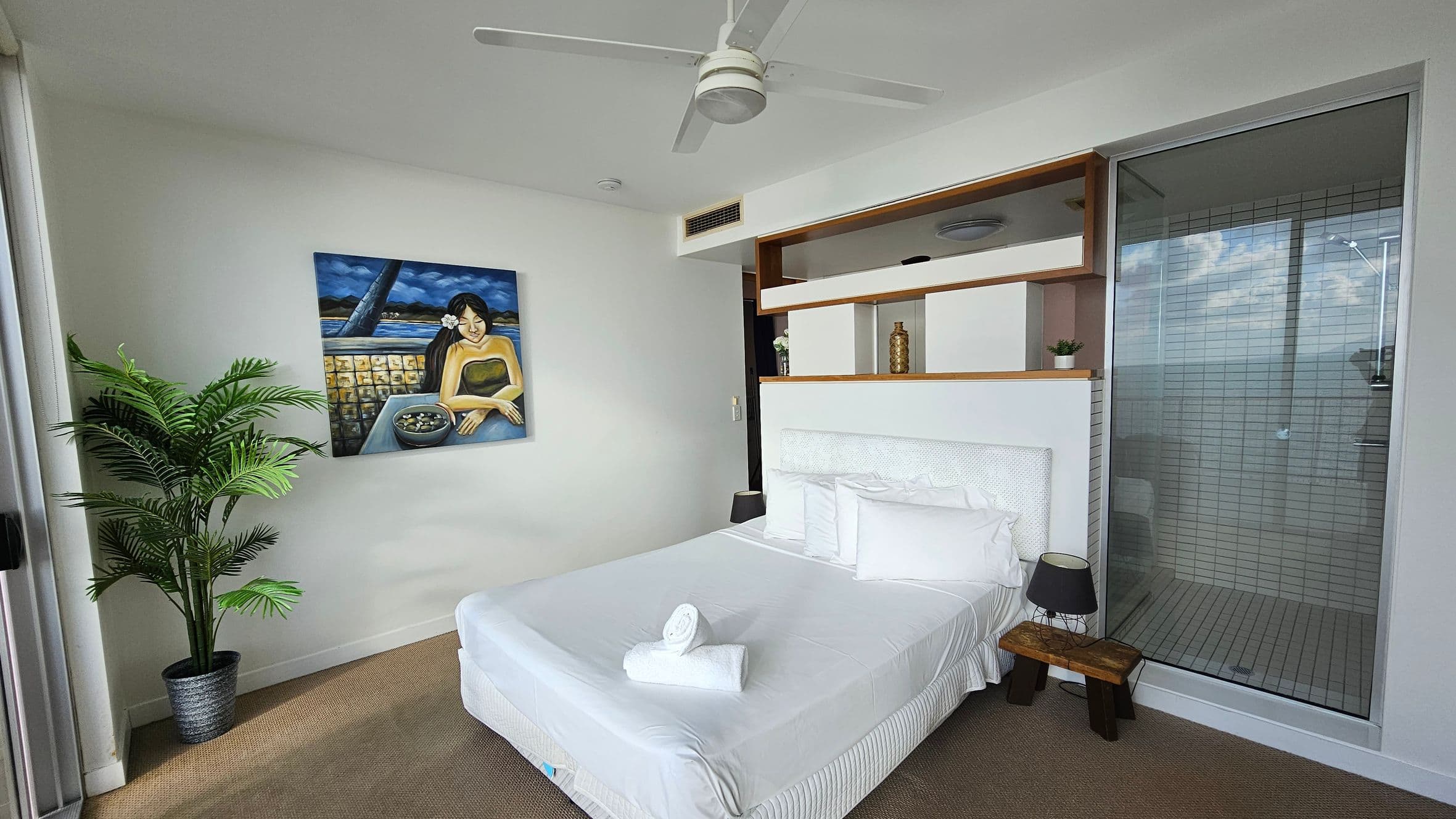 A bedroom with a white bed in the centre, topped with neatly arranged pillows and a rolled towel. To the left, a potted plant stands beside a painting of a woman at a table. On the right, a glass-walled shower area is visible. Above the bed, a ceiling fan is mounted. The room features a shelf with decorative items and a small lamp on a wooden stool beside the bed. The floor is carpeted in a neutral tone.