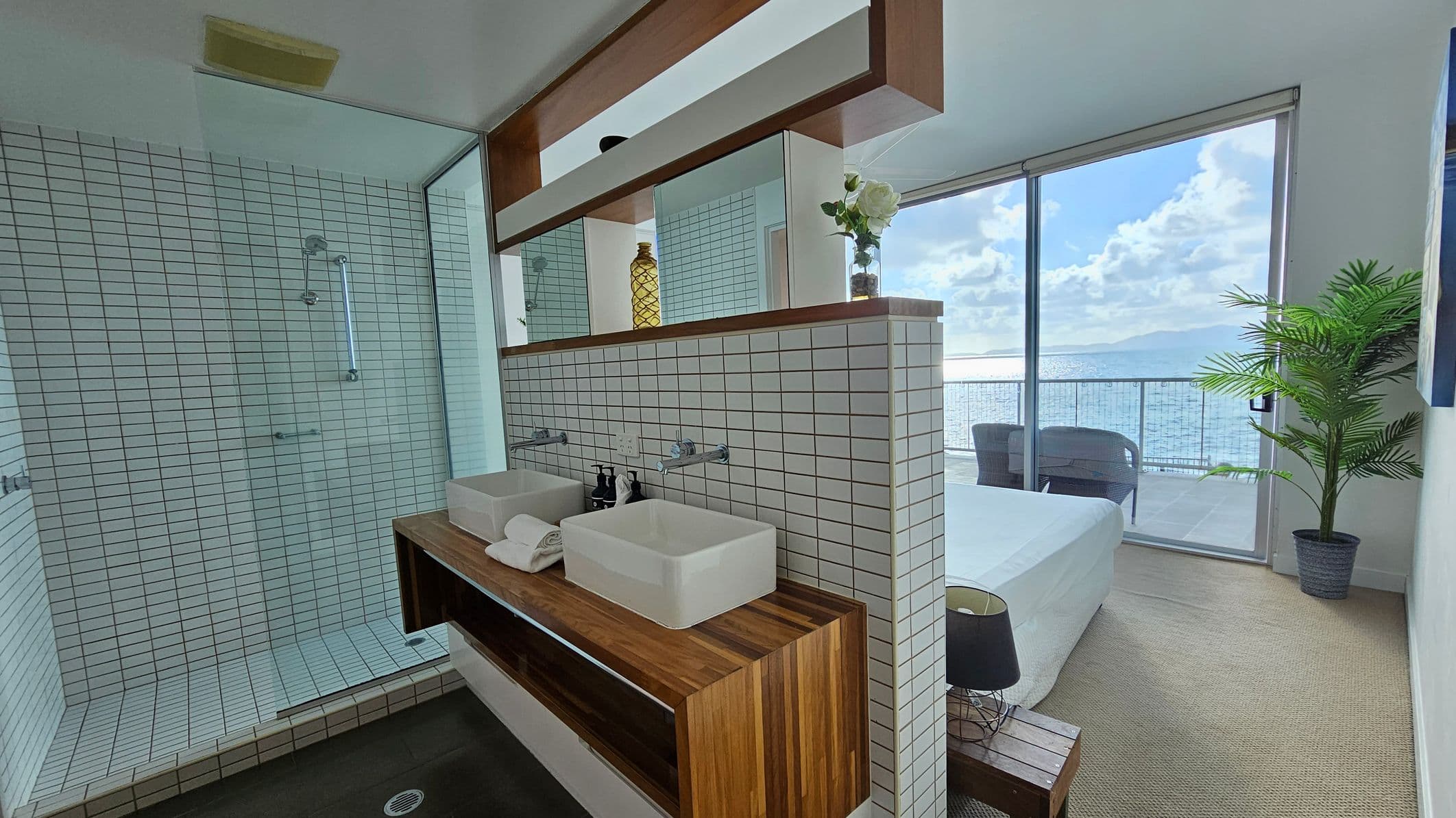 A modern bathroom and bedroom with a view. On the left, there's a glass-enclosed shower with white tiles. In the centre, a wooden vanity with two square sinks and a mirrored shelf above. On the right, a bedroom with a bed, a small bedside table with a lamp, and a potted plant. Large glass doors open to a balcony with chairs, overlooking the sea. The floor is carpeted, and the ceiling is plain white.