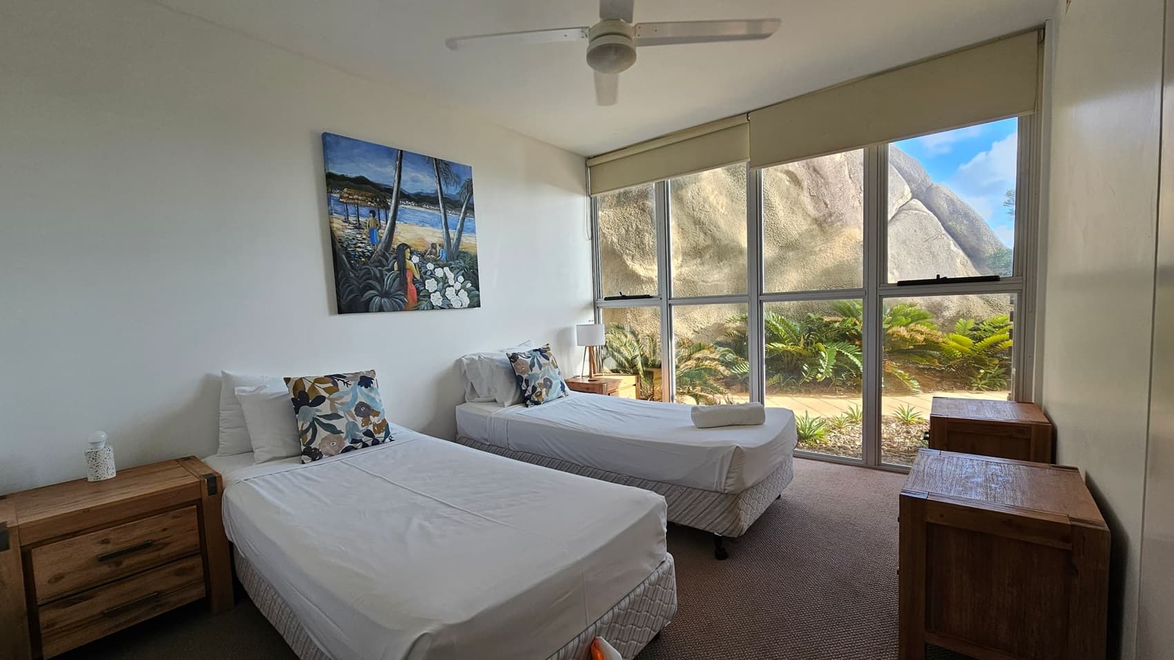 A bedroom with two single beds, each with white bedding and floral cushions. A wooden bedside table is next to each bed, with a lamp on the right table. A colourful painting hangs above the left bed. Large windows on the right reveal a view of rocks and greenery outside. The room has a ceiling fan and a neutral carpeted floor.