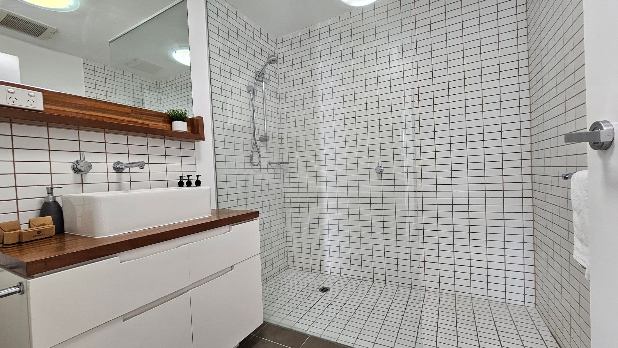 A modern bathroom with white square tiles covering the walls and floor. On the left, there's a wooden countertop with a white rectangular sink, a soap dispenser, and small bottles. Above the sink is a wooden shelf with a small potted plant. The shower area is on the right, featuring a handheld showerhead and a glass partition. A white towel hangs on a rail attached to the open door. The ceiling has recessed lighting.