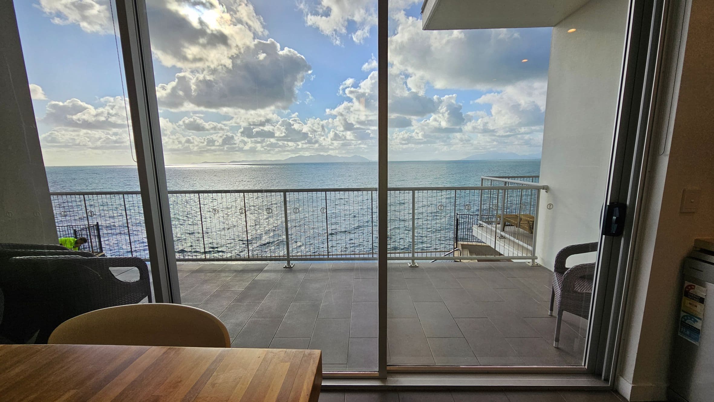 A view through a large glass sliding door showing a balcony with a metal railing overlooking the sea. The balcony has grey tiles and a wicker chair on the right. Inside, a wooden table is partially visible at the bottom, with a chair on the left. The horizon shows distant landforms under a sky filled with clouds.