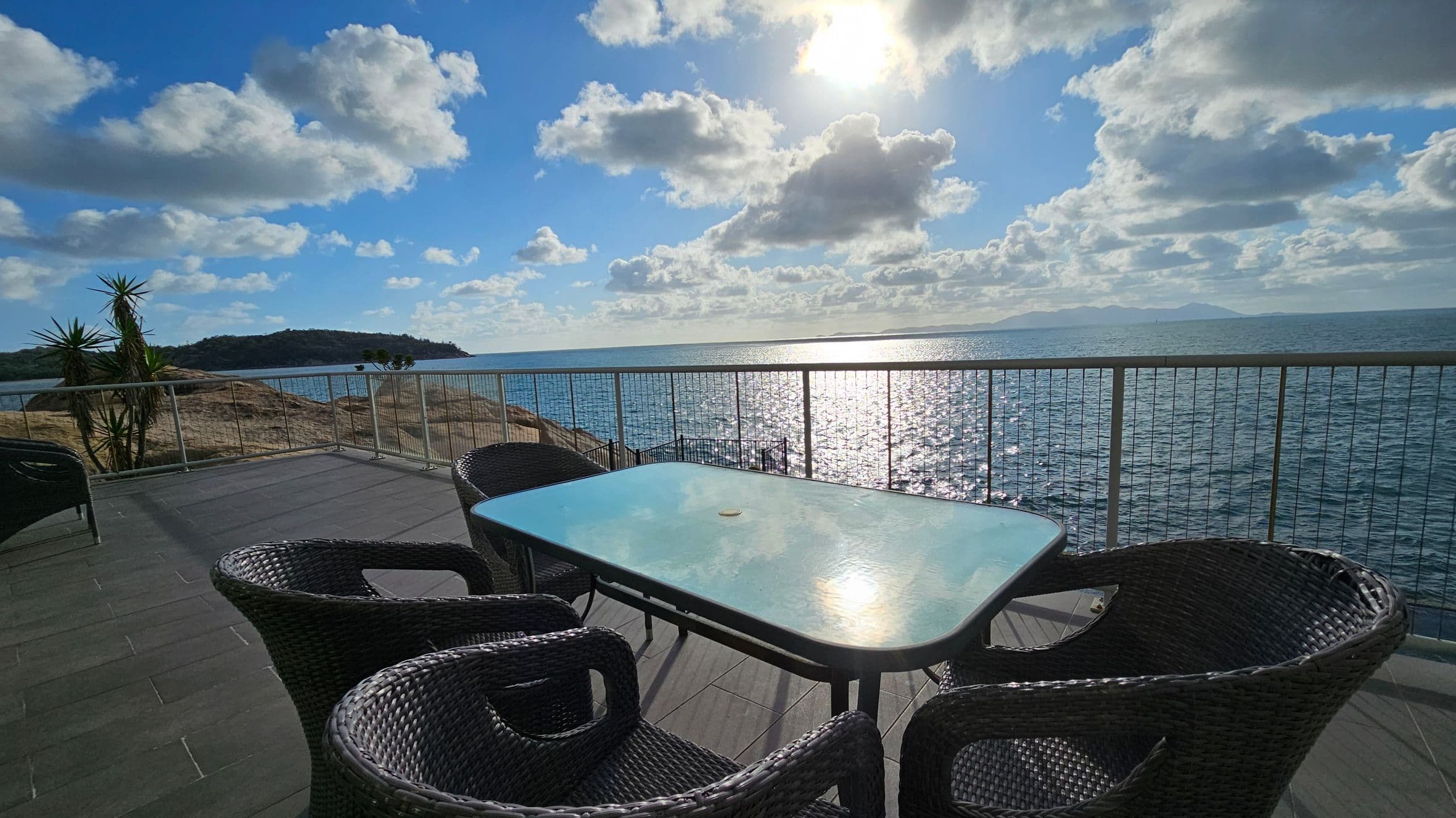 A terrace with a glass table and four wicker chairs overlooks a vast expanse of water. The sun reflects off the sea, creating a shimmering effect. To the left, there are a few tall plants and a rocky outcrop. The terrace has a tiled floor and is bordered by a metal railing. In the distance, a small island is visible under a partly cloudy sky.