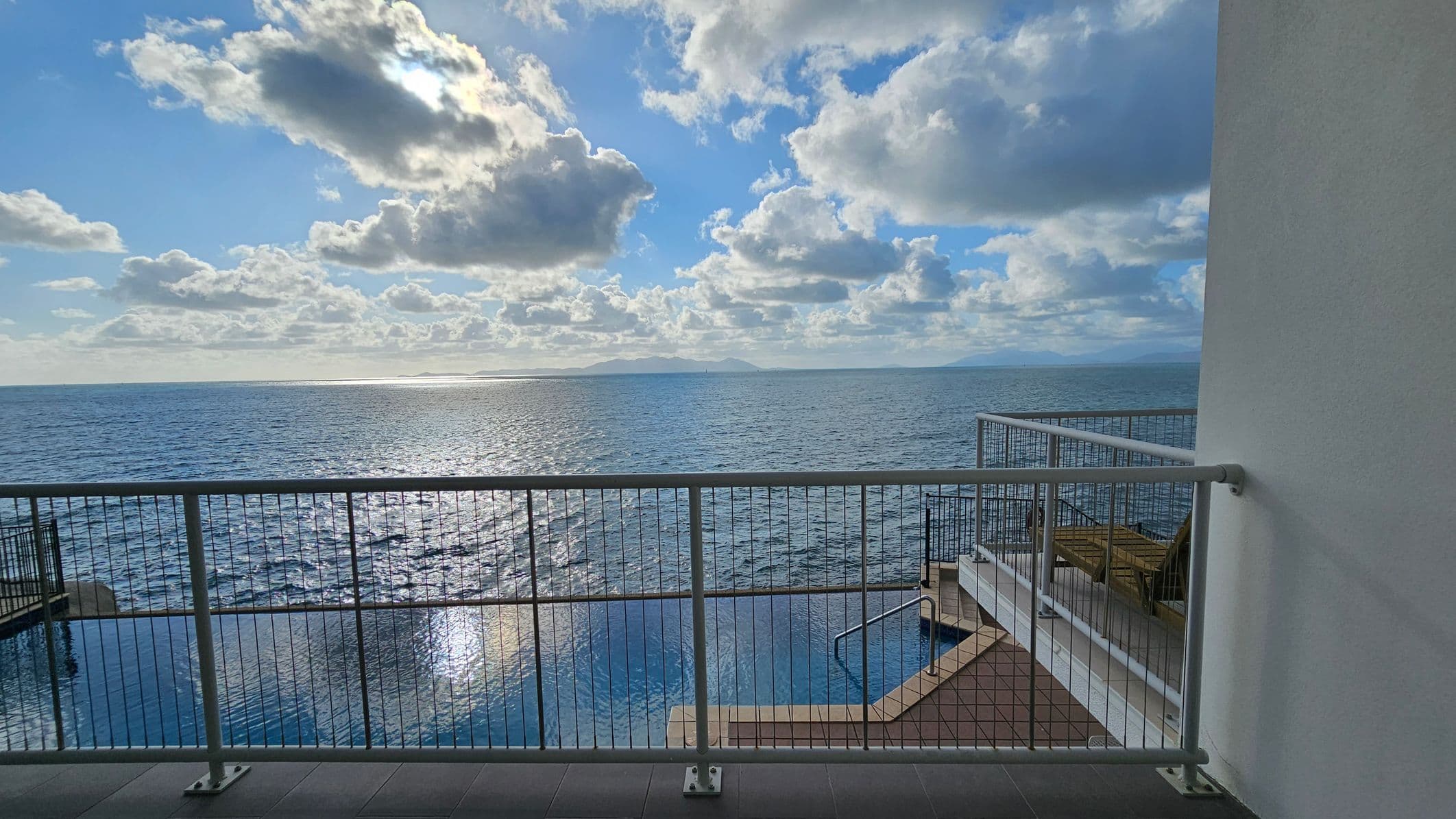 A view from a balcony overlooking a vast body of water. The foreground features a metal railing, with a tiled floor beneath. To the right, there is a section of a building wall. Below the balcony, a swimming pool is visible, with steps leading into it. In the distance, the horizon meets the water, with land faintly visible. The sky is filled with scattered clouds, and sunlight reflects off the water's surface.