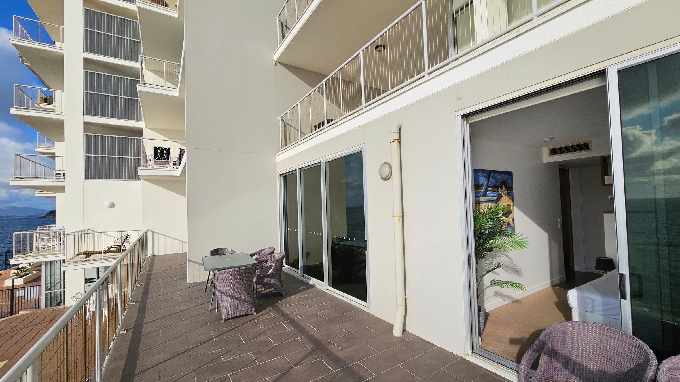 A spacious balcony with brown tiled flooring and a glass railing overlooks the ocean. On the right, a set of wicker chairs and a table are placed near large sliding glass doors. Inside, a room is visible with a painting on the wall and a potted plant. The building's exterior is light-coloured with multiple balconies on the left side. The ocean is visible in the background.