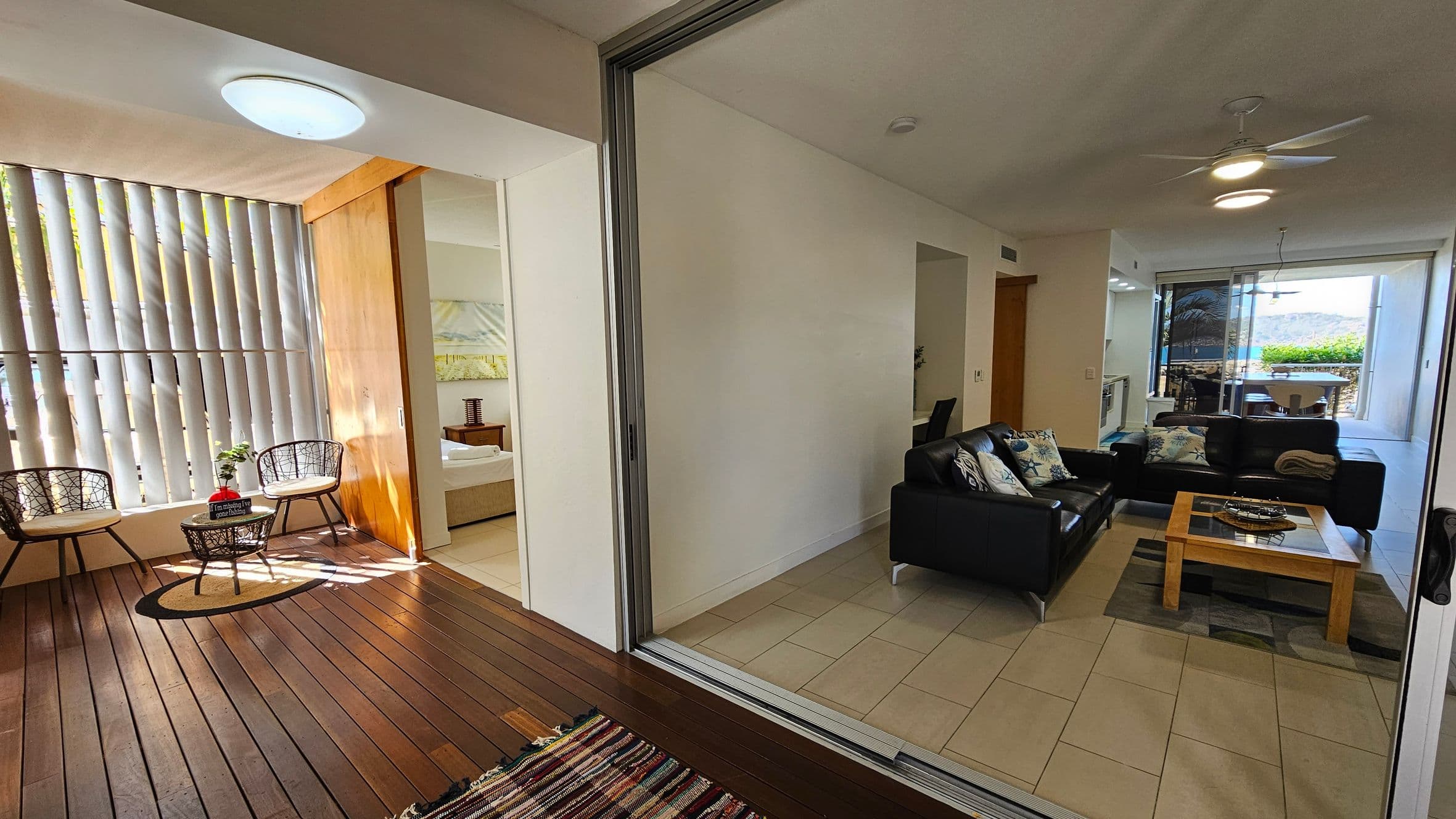 A modern interior with a wooden-floored area on the left featuring two wicker chairs and a small table with a plant. Vertical blinds cover the window. Adjacent is a bedroom with a visible bed. On the right, a tiled living room has a black leather sofa set with patterned cushions and a wooden coffee table. A ceiling fan is above. In the background, a dining area with a view of greenery and water is visible through large windows.