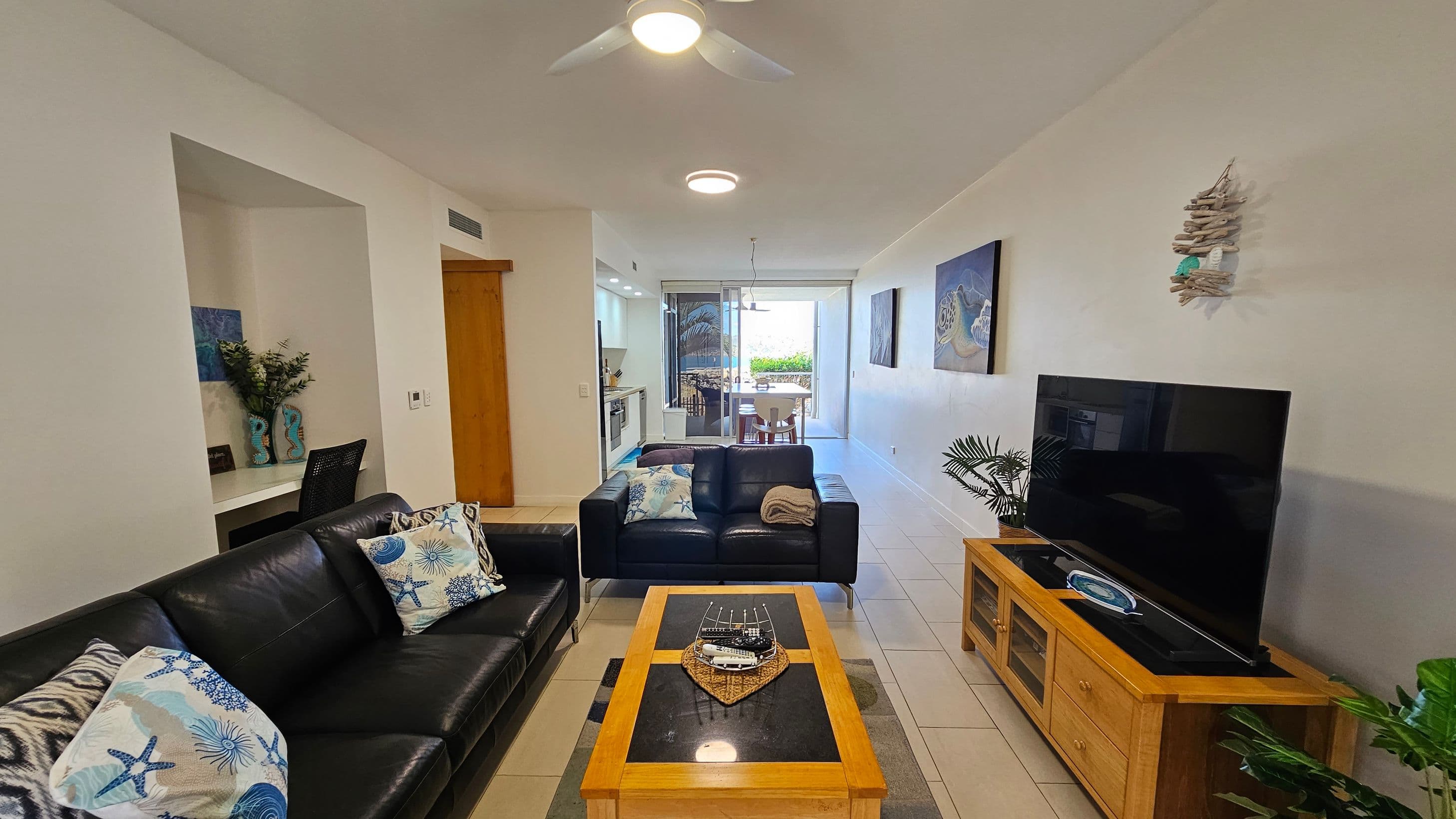 A modern living room with a black leather sofa adorned with patterned cushions on the left. In the centre, a wooden coffee table with a decorative tray. On the right, a large flat-screen TV sits on a wooden stand with plants nearby. The background features a dining area with a table and chairs, and a kitchen with visible appliances. The walls are decorated with artwork, and the ceiling has recessed lighting and a fan. The floor is tiled.