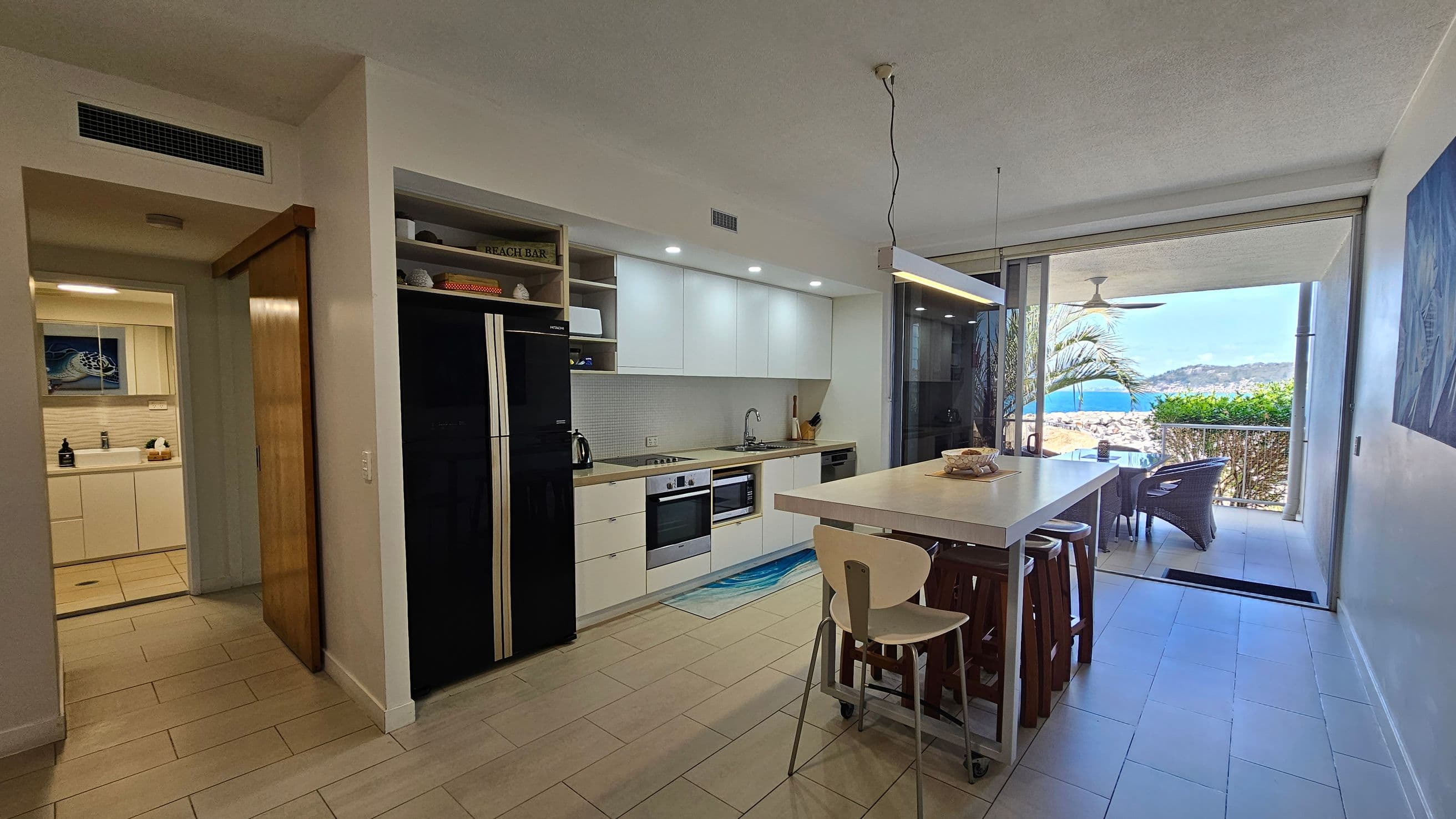 Modern kitchen with light-coloured cabinets and a black fridge on the left. A wooden sliding door leads to a bathroom with a sink and artwork. In the centre, a white island with stools and a hanging light fixture. On the right, large glass doors open to a balcony with wicker chairs and a view of the sea and hills. The floor is tiled, and the ceiling is smooth with recessed lighting.
