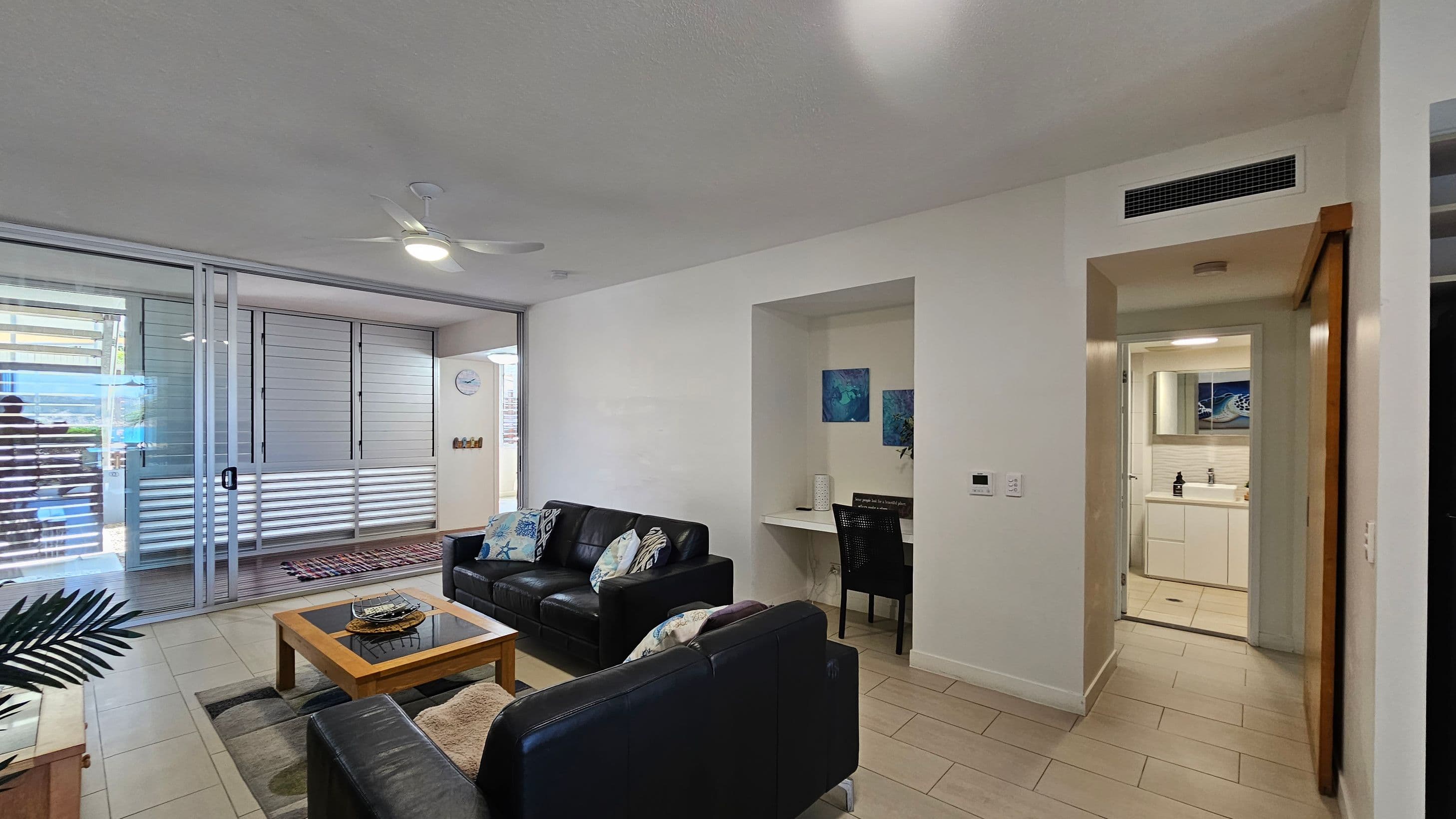 A modern living room with a tiled floor and a ceiling fan. On the left, there are sliding glass doors with horizontal blinds leading to an outdoor area. In the centre, two black leather sofas with patterned cushions face a wooden coffee table. On the right, a small alcove with a desk and chair is visible, along with a doorway leading to a bathroom with a white vanity and mirror. A potted plant is partially visible in the foreground.