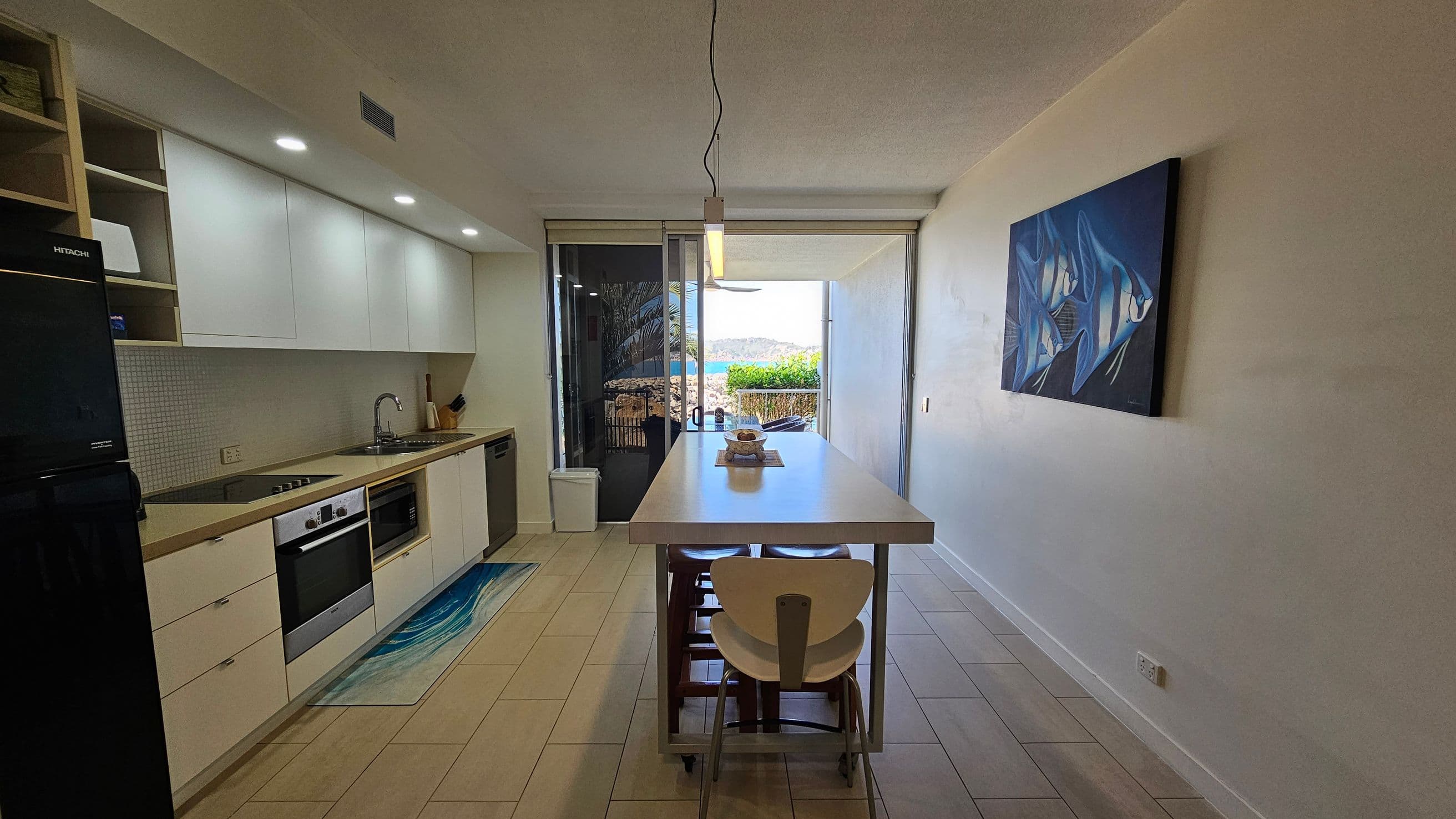 A modern kitchen with light-coloured cabinets and a black fridge on the left. The countertop features a sink and an oven. A long dining table with chairs is in the centre, with a hanging light above. On the right wall, there's a painting of fish. At the far end, glass doors open to a view of greenery and water. The floor is tiled, and the ceiling is plain with recessed lighting.