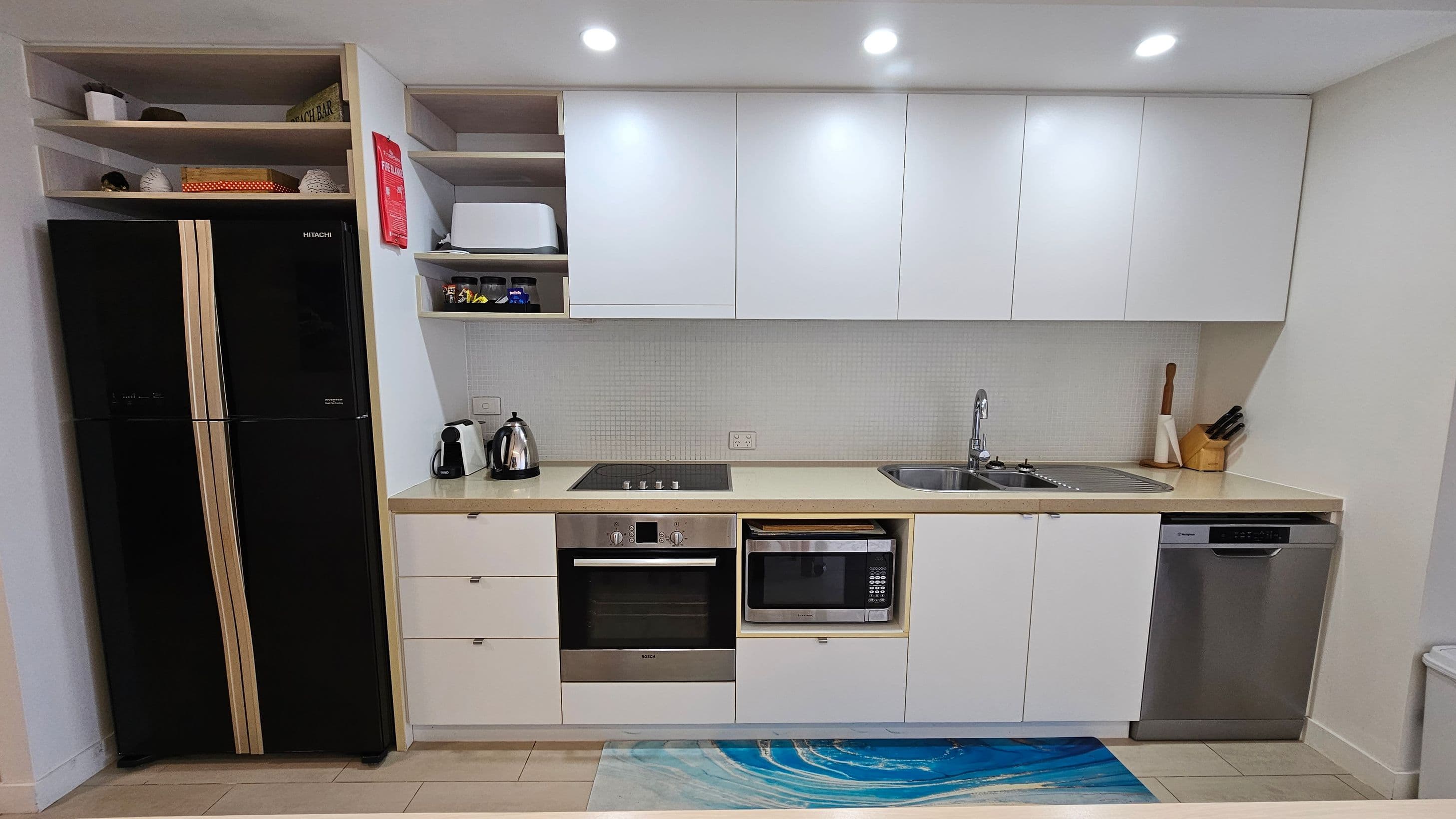 A modern kitchen with a black double-door fridge on the left, surrounded by open shelves holding decorative items. Next to it is a countertop with a kettle and coffee machine, and a built-in oven and microwave. Above are white cabinets. The sink is on the right, with a knife block and cutting board nearby. Below the sink is a dishwasher. The floor is tiled with a blue patterned mat in front of the counter. The ceiling has recessed lighting.