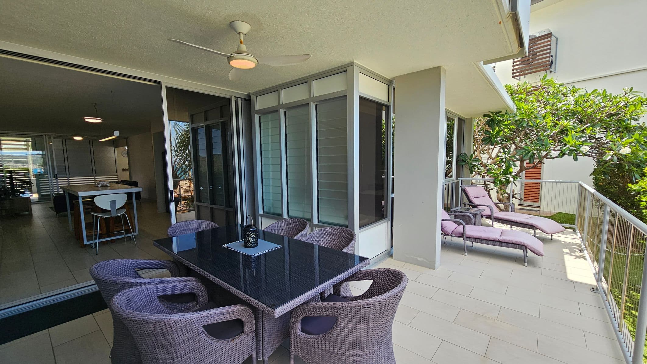A modern outdoor patio with a glass-top dining table and six wicker chairs on the left. A ceiling fan is above. On the right, two cushioned sun loungers are positioned near a railing, with a leafy tree providing some shade. The floor is tiled, and large sliding glass doors lead to an interior space with a kitchen island and bar stool visible.