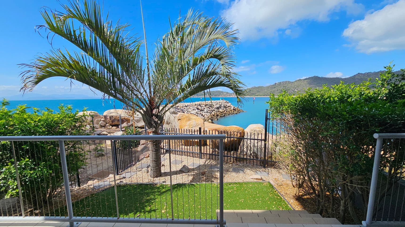 A view from a patio with a metal railing in the foreground. A palm tree stands prominently in the centre, with a small patch of grass and stone pathway beneath it. Beyond the railing, there is a rocky shoreline leading to a calm blue sea. On the right, dense green bushes frame the scene. In the background, a breakwater extends into the water, and hills are visible in the distance under a partly cloudy sky.
