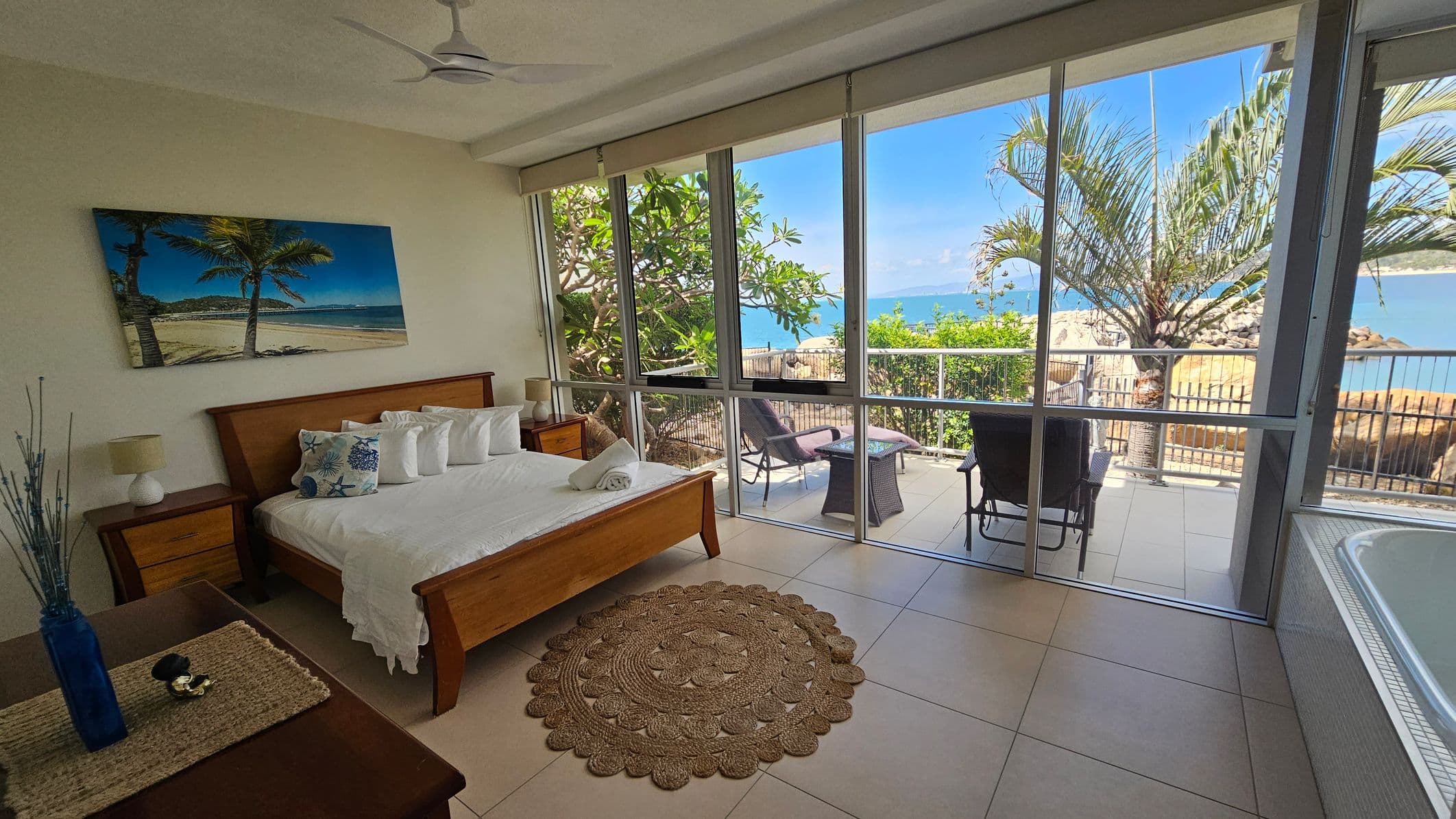 A bedroom with a large window view of the ocean. On the left, there's a wooden bed with white and patterned pillows, flanked by two bedside tables with lamps. A beach-themed painting hangs above the bed. In the middle, a round woven rug lies on the tiled floor. On the right, glass doors open to a patio with chairs and a table, surrounded by greenery and a palm tree. A ceiling fan is visible above.