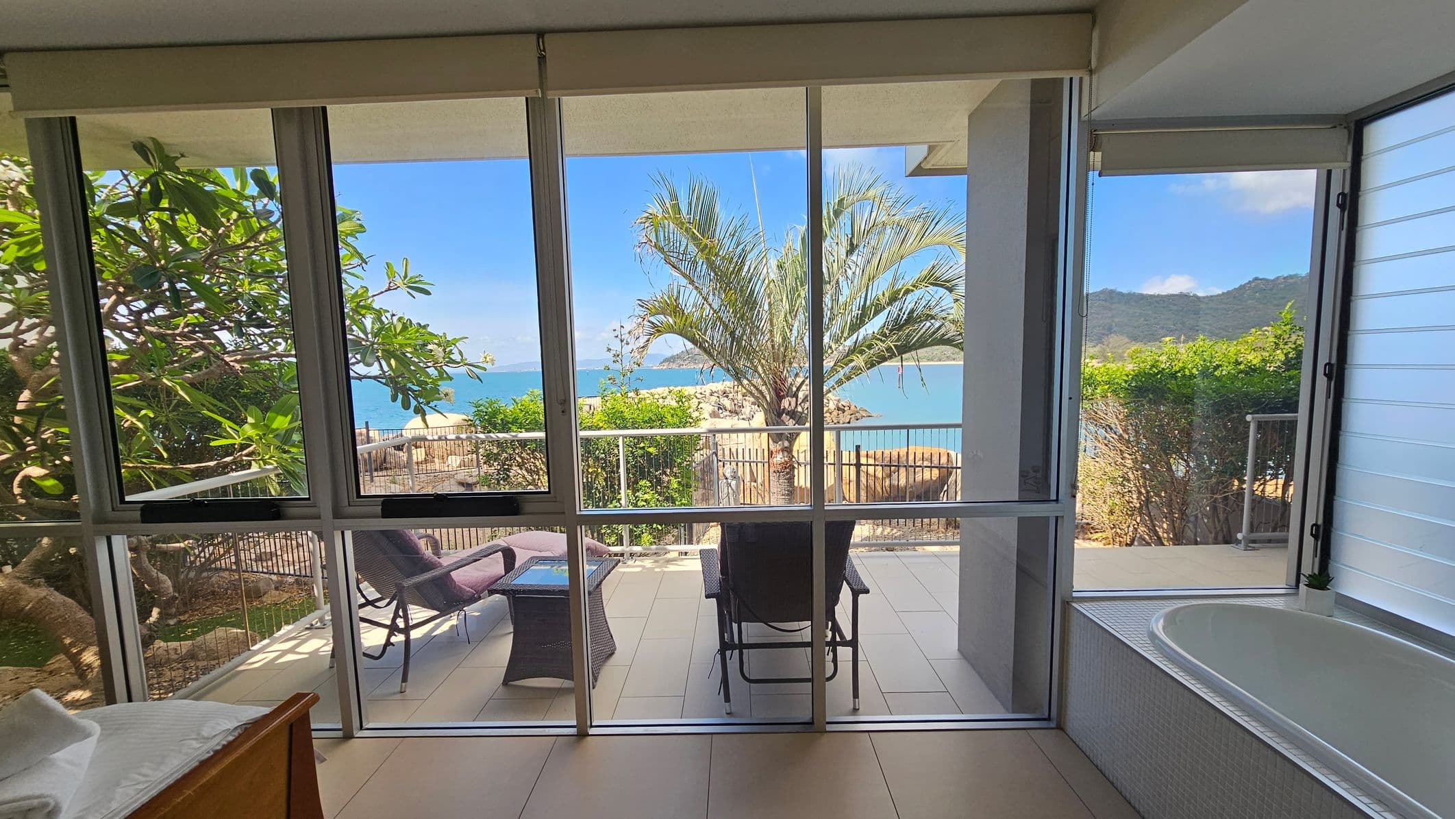 A room with large glass windows overlooking a patio. On the left, there's a leafy tree visible through the window. In the centre, the patio has a chair and a small table, with a view of the ocean and a palm tree. On the right, a bathtub is positioned near the window. The background shows a clear blue sea and distant hills. The floor is tiled, and the ceiling is not visible.