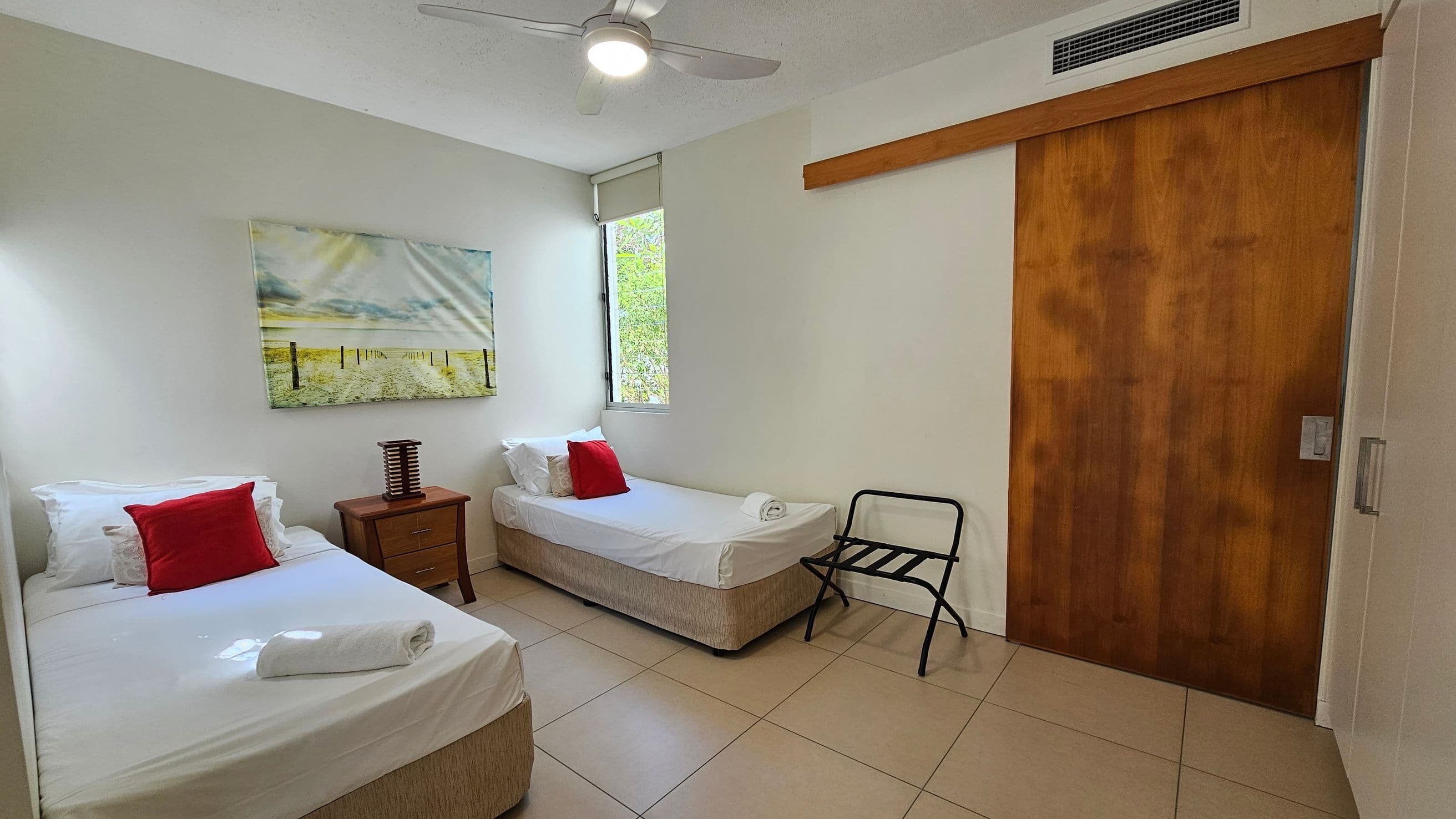 A small bedroom with two single beds, each with white sheets and a red cushion. A rolled towel is placed on each bed. Between the beds is a wooden nightstand with a decorative lamp. Above the left bed is a landscape painting. A window with a blind is on the back wall. A wooden sliding door is on the right, next to a black luggage rack. The floor is tiled, and a ceiling fan with a light is above.