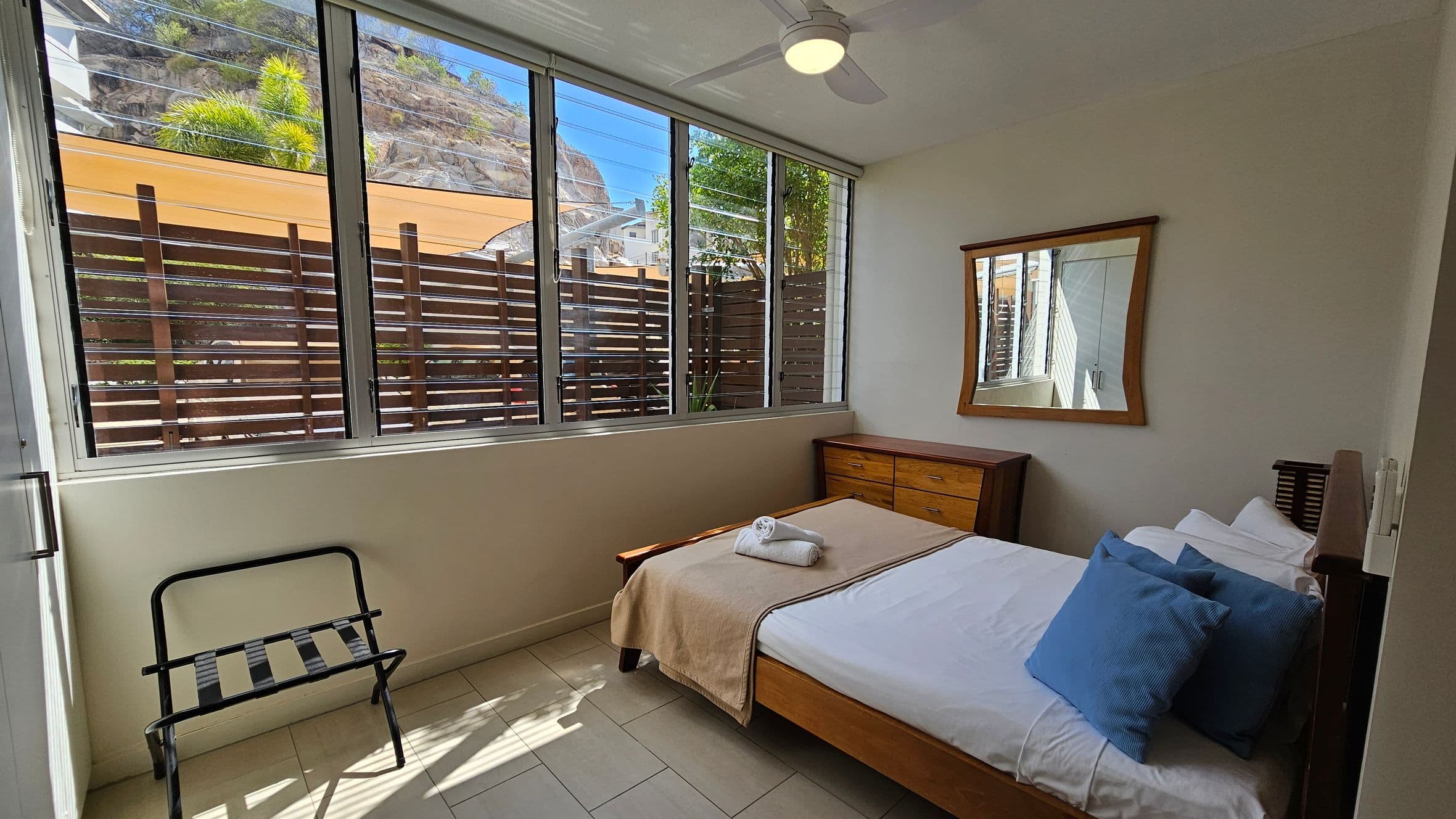 A small bedroom with a bed on the right, covered with a beige blanket and blue cushions. A rolled towel is placed on the bed. Above the bed is a wooden-framed mirror. A wooden chest of drawers is next to the bed. On the left, a luggage rack sits near a large window with horizontal blinds, allowing natural light to enter. Outside, there is a wooden fence and some greenery. The floor is tiled, and a ceiling fan is visible above.