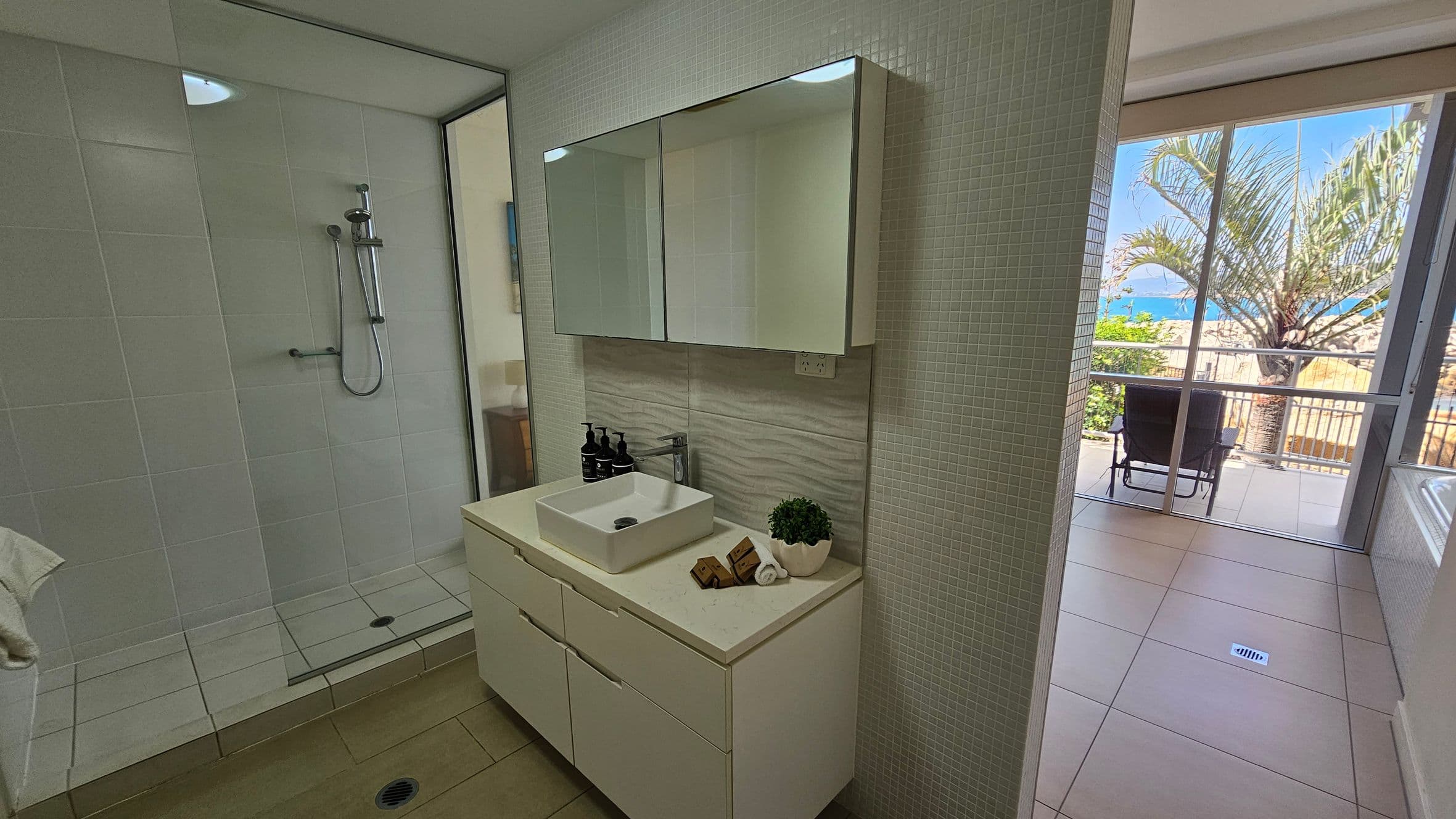 On the left, a bathroom with a glass-enclosed shower featuring a modern showerhead and white tiles. In the middle, a white vanity with a square basin, a mirror cabinet above, and toiletries on the counter. On the right, a doorway leads to a balcony with a view of a palm tree and the sea. The floor is tiled, and the balcony has a chair facing the ocean.