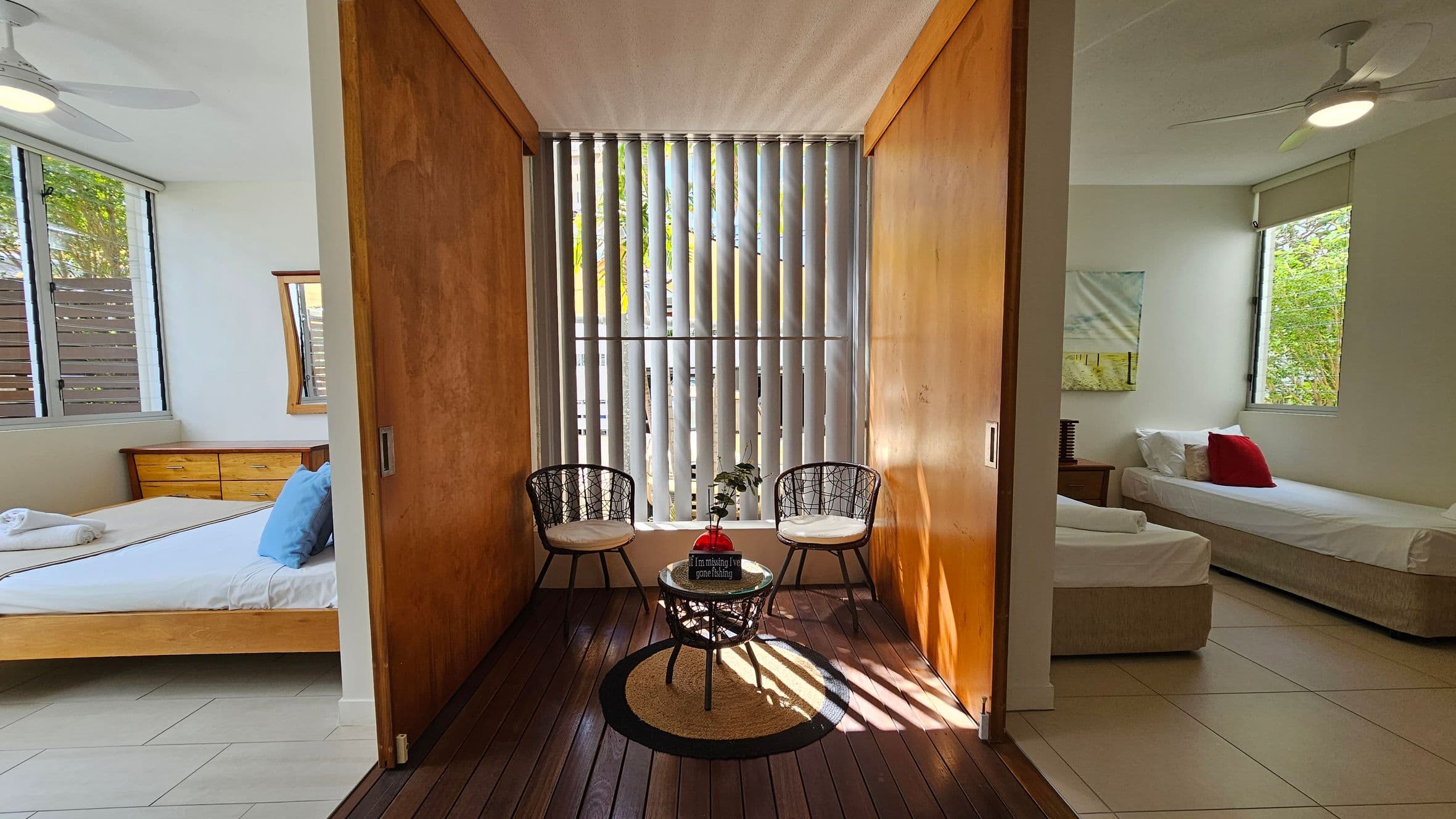A room with two sections divided by wooden sliding doors. On the left, there's a bedroom with a wooden bed, white bedding, and a wooden dresser. On the right, another room with a single bed, white bedding, and a red cushion. In the centre, a small seating area with two black wire chairs, a round table, and a plant in a red vase. Vertical blinds cover the window, and the floor is a mix of wood and tiles. Ceiling fans are visible in both sections.