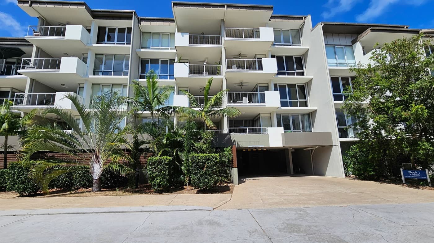 A modern residential complex with multiple buildings featuring large windows and balconies. In the foreground, a curved road leads to the complex, with a landscaped area containing a tall palm tree and a "Shared Zone" sign. On the right, there is a covered parking area with several parked cars. The background includes a rocky hillside with greenery. The sky is clear and blue.
